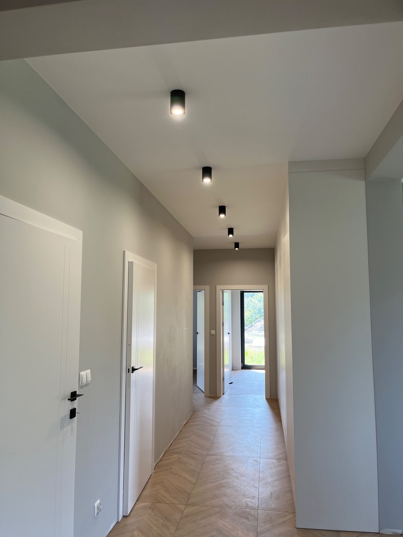 Korytarz w jasnych barwach z parkietem ułożonym we wzór jodełki; na suficie zamontowane czarne, punktowe oprawy oświetleniowe.