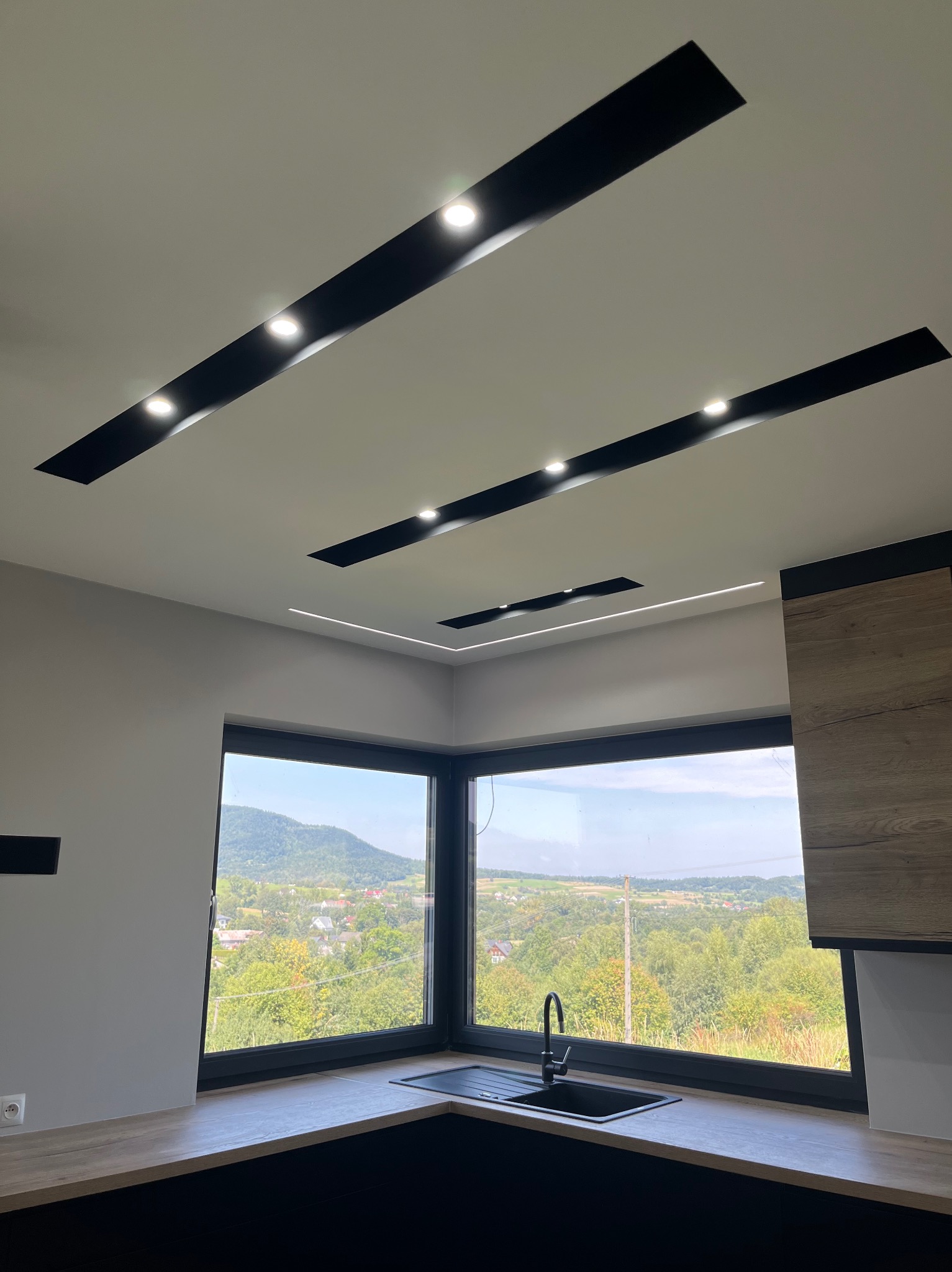 Nowoczesna kuchnia z oknem narożnym i widokiem na zielone wzgórza, oświetlenie sufitowe w postaci czarnych listew z punktowymi lampami oraz liniowe oświetlenie LED w rogu sufitu.