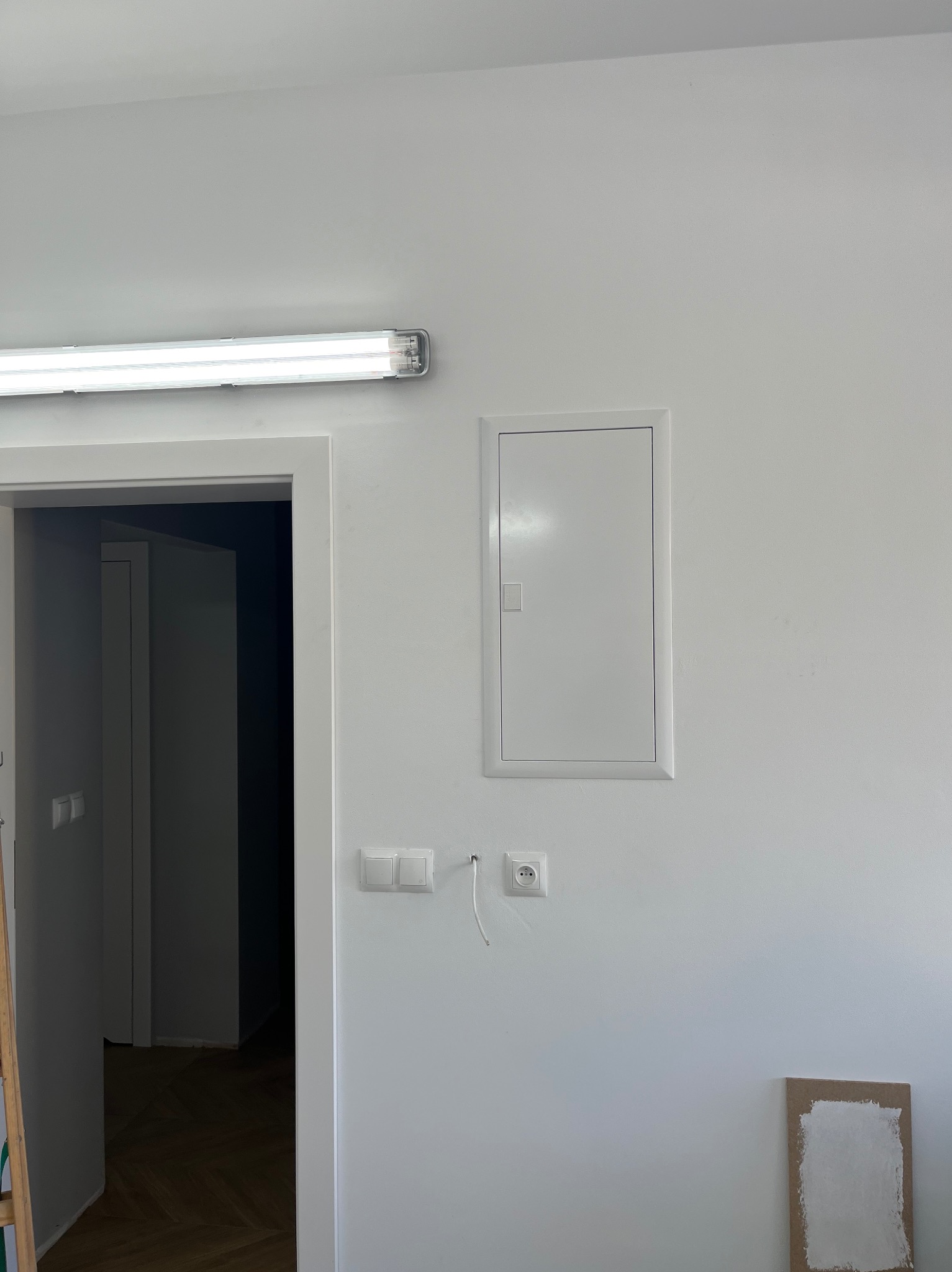 Biała ściana z zamontowaną skrzynką elektryczną, podwójnym włącznikiem światła i gniazdkiem z wystającym przewodem. Nad drzwiami lampa fluorescencyjna. Na podłodze karton z próbką białej farby.