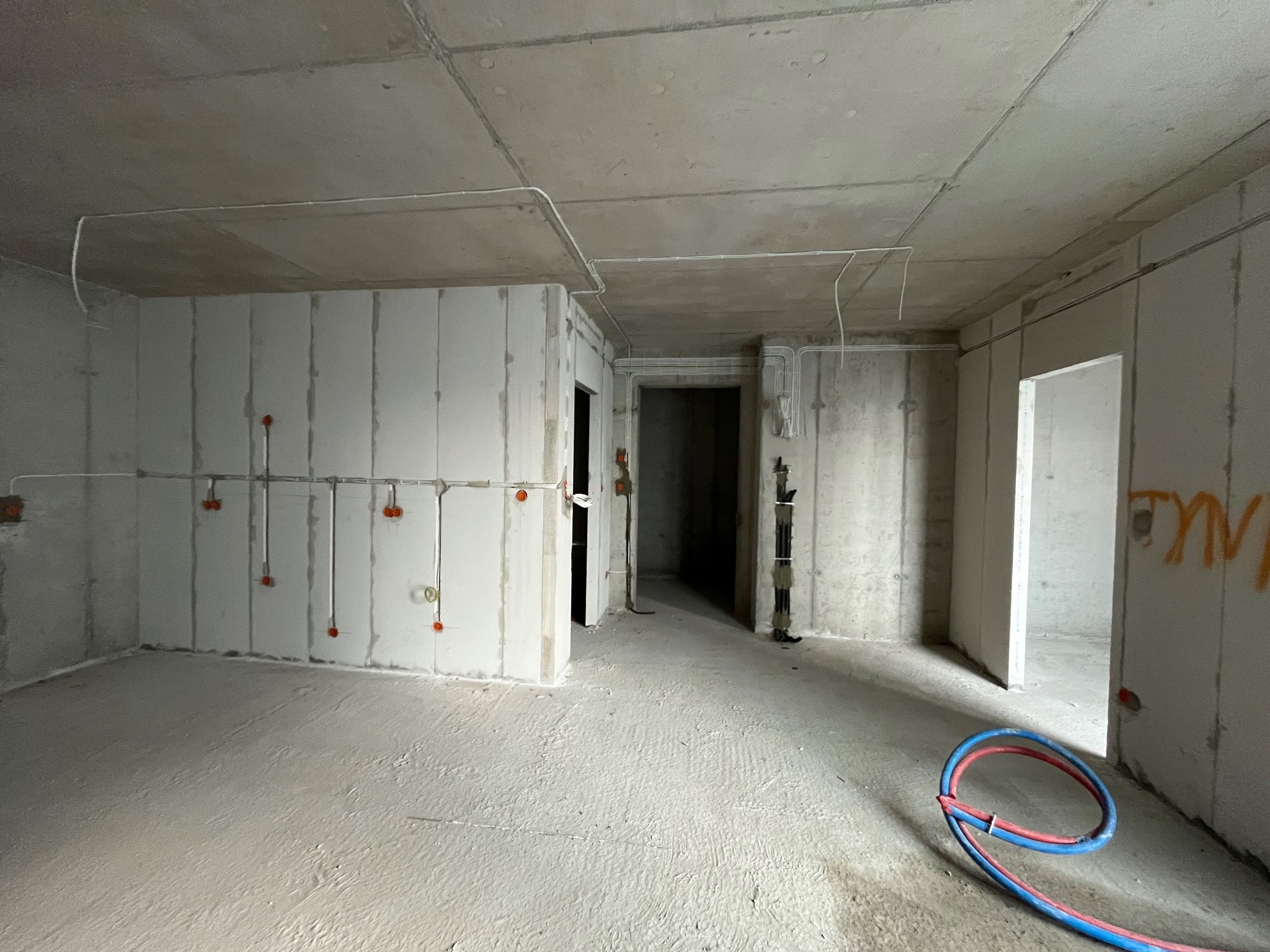 Surowy stan wnętrza budynku w trakcie prac elektrycznych: widoczne kable prowadzone po suficie i ścianach, puszki instalacyjne na ścianie oraz zwój niebiesko-czerwonego przewodu na podłodze.