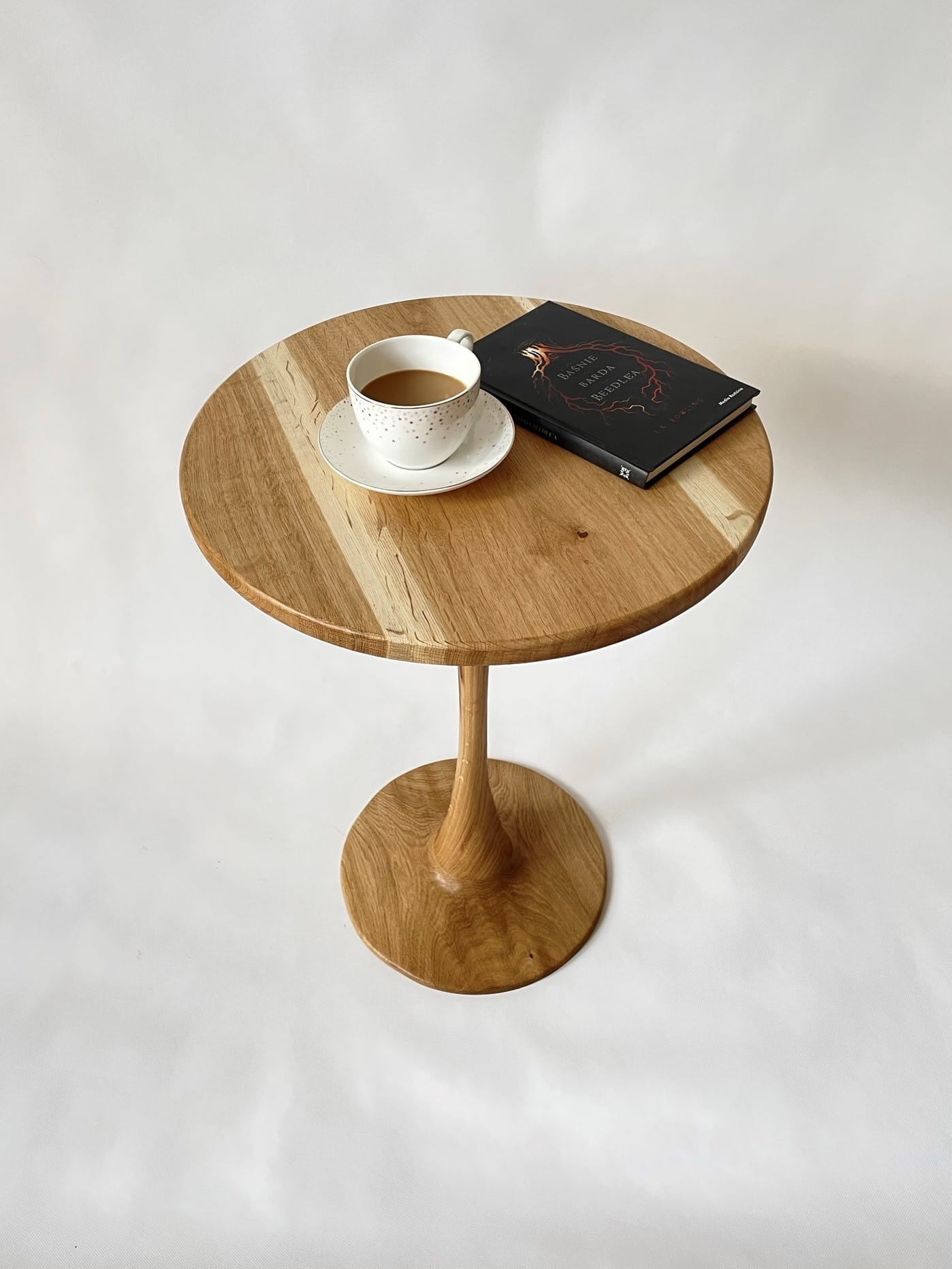 Okrągły, drewniany stolik kawowy z filiżanką kawy i książką na blacie, wykończony w jasnym odcieniu drewna dębowego, na białym tle.