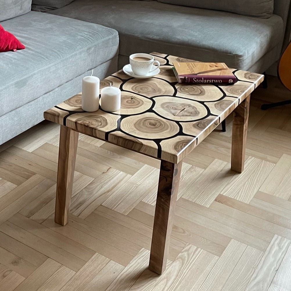 Stolik kawowy z blatem z plastrów drewna zalanych żywicą, na blacie książka o stolarstwie, filiżanka kawy i dwie białe świece, w tle szara sofa.
