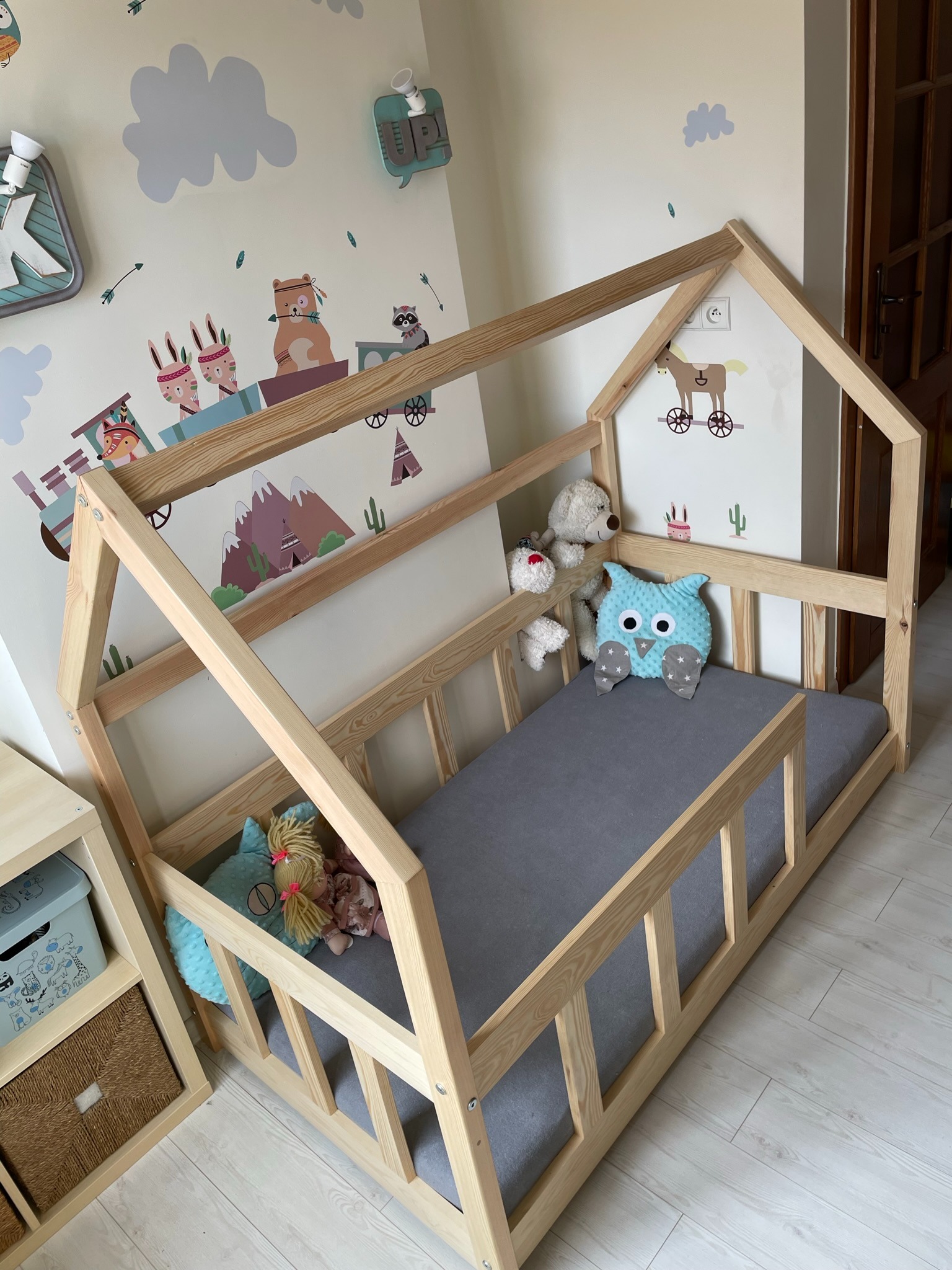 Drewniane łóżko dziecięce w kształcie domku z szarym materacem, pluszowymi zabawkami i poduszką w kształcie sowy, na tle ściany z naklejkami w stylu indiańskim.