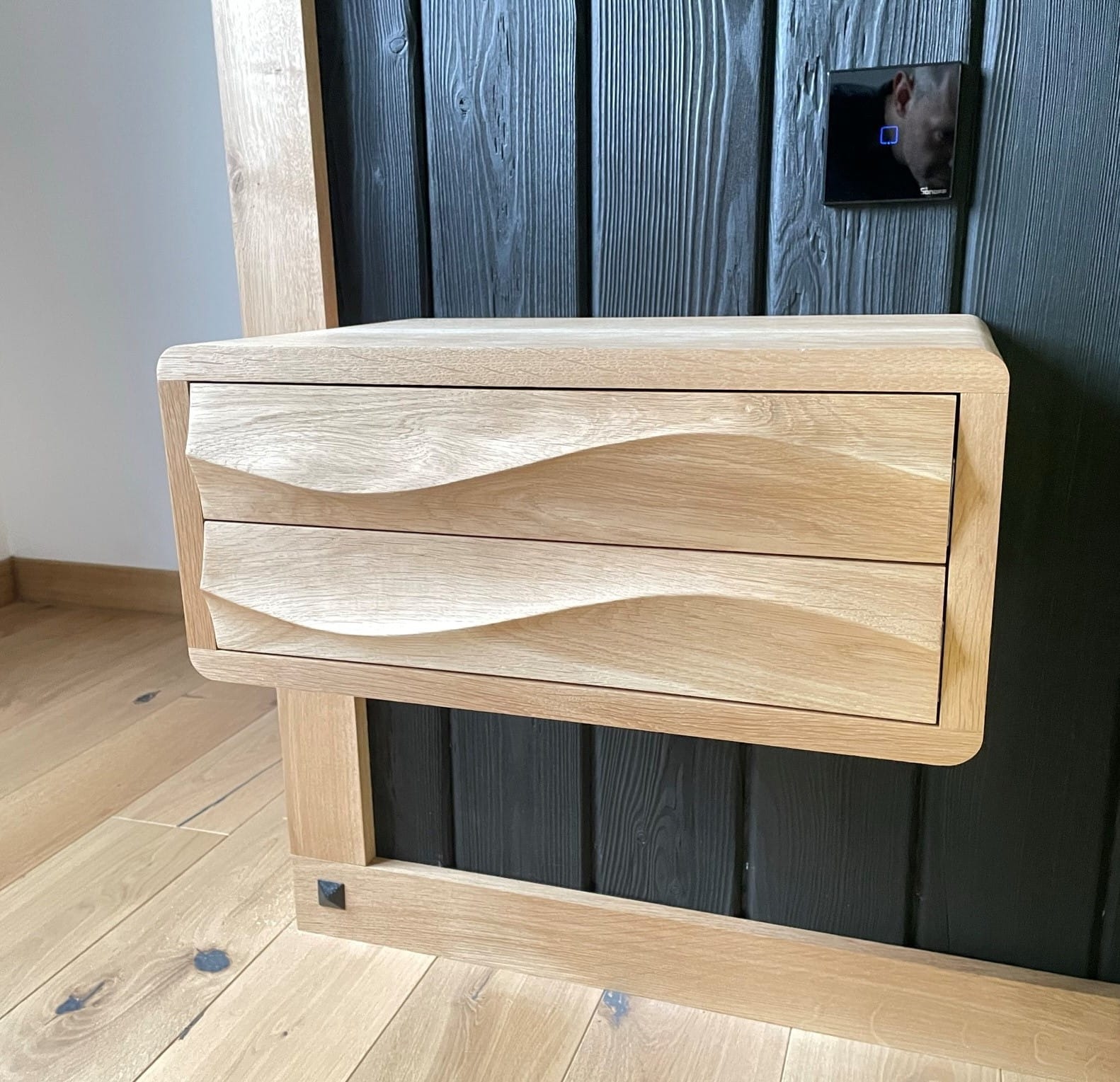 Drewniana szafka nocna z dwoma szufladami o falistych frontach, przymocowana do czarnej, drewnianej ściany z widocznym włącznikiem dotykowym, na tle drewnianej podłogi.