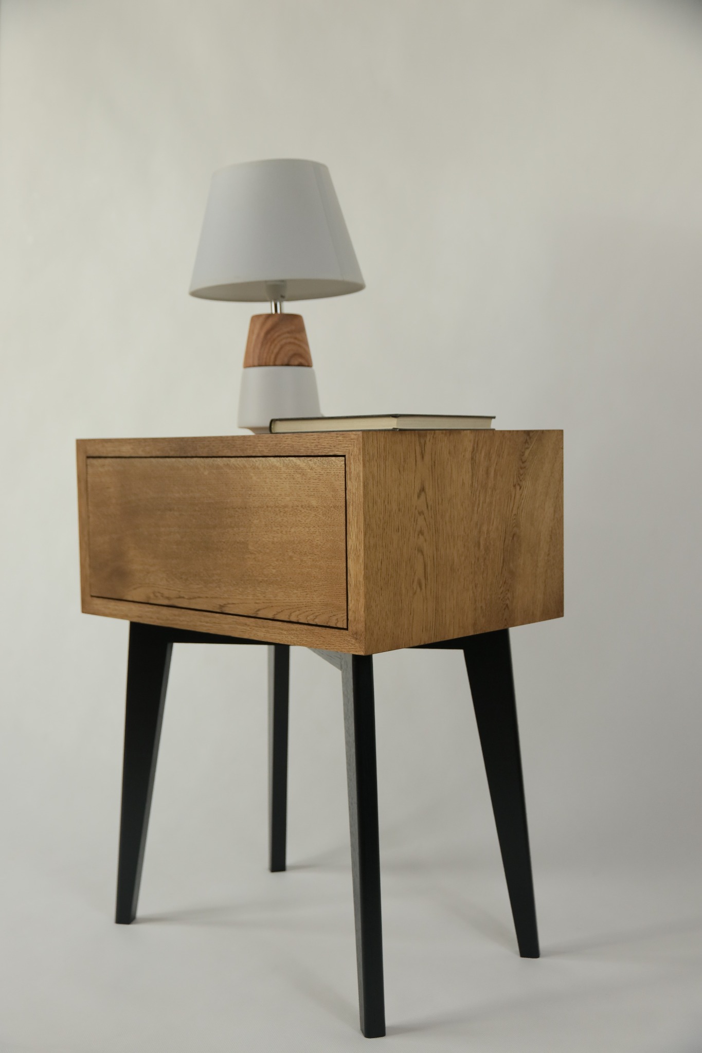 Drewniana szafka nocna z szufladą i stożkowymi czarnymi nogami, na blacie leży książka, a obok stoi lampka nocna z białym abażurem i drewnianą podstawą, na jasnym tle.