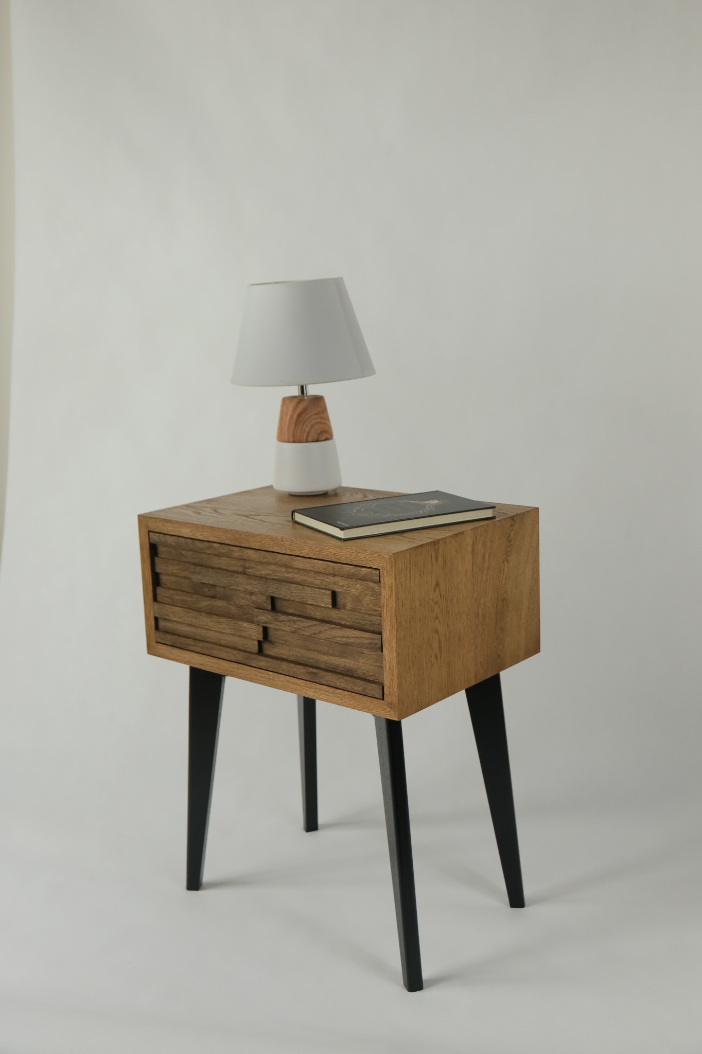 Drewniana szafka nocna z trzema szufladami o unikalnym wzorze frontów, stojąca na czarnych, smukłych nogach, z lampką nocną i książką na blacie na neutralnym tle.