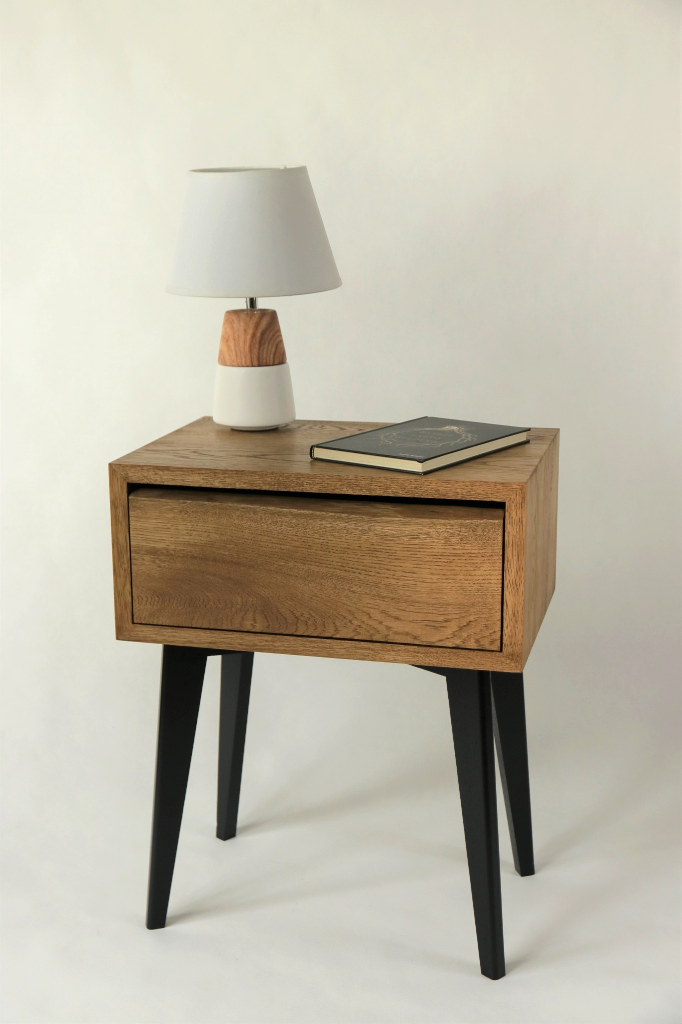 Drewniana szafka nocna z pojedynczą szufladą, czarne nóżki, na blacie lampka z drewnianą podstawą i książka w ciemnej okładce, neutralne tło.