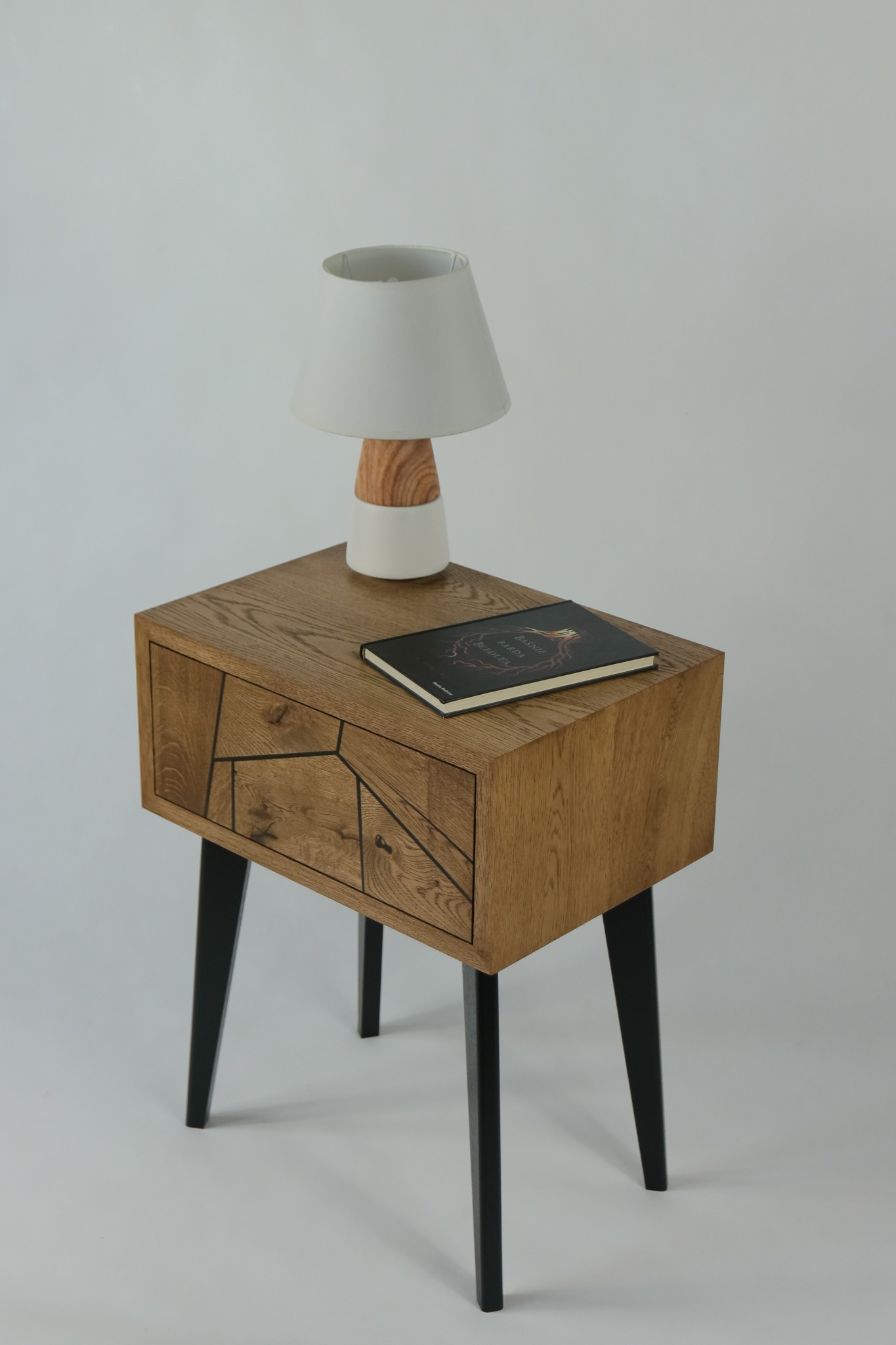 Drewniana szafka nocna z geometrycznym wzorem na szufladzie, czarne nóżki, na blacie leży książka i stoi lampka z drewnianą podstawą na białym tle.