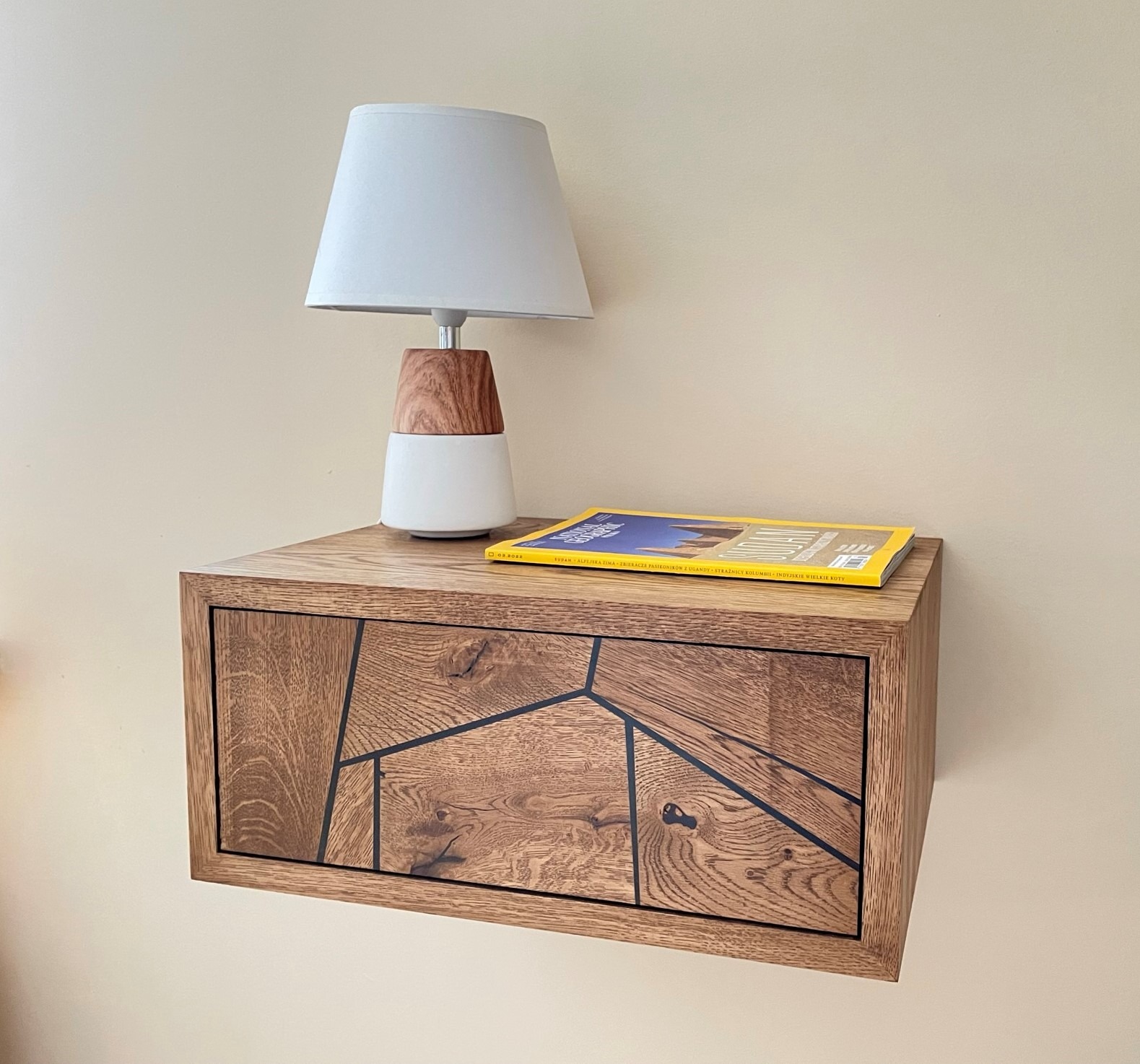 Drewniana szafka nocna wisząca z geometrycznym wzorem na froncie szuflady, na niej lampka nocna z abażurem i magazyn.