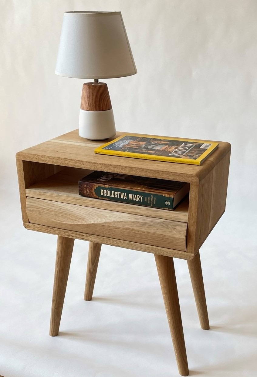 Drewniana szafka nocna z szufladą i półką, na której leży książka 'Królestwa Wiary' i magazyn National Geographic, a na blacie stoi lampka nocna z drewnianą podstawą i beżowym abażurem.