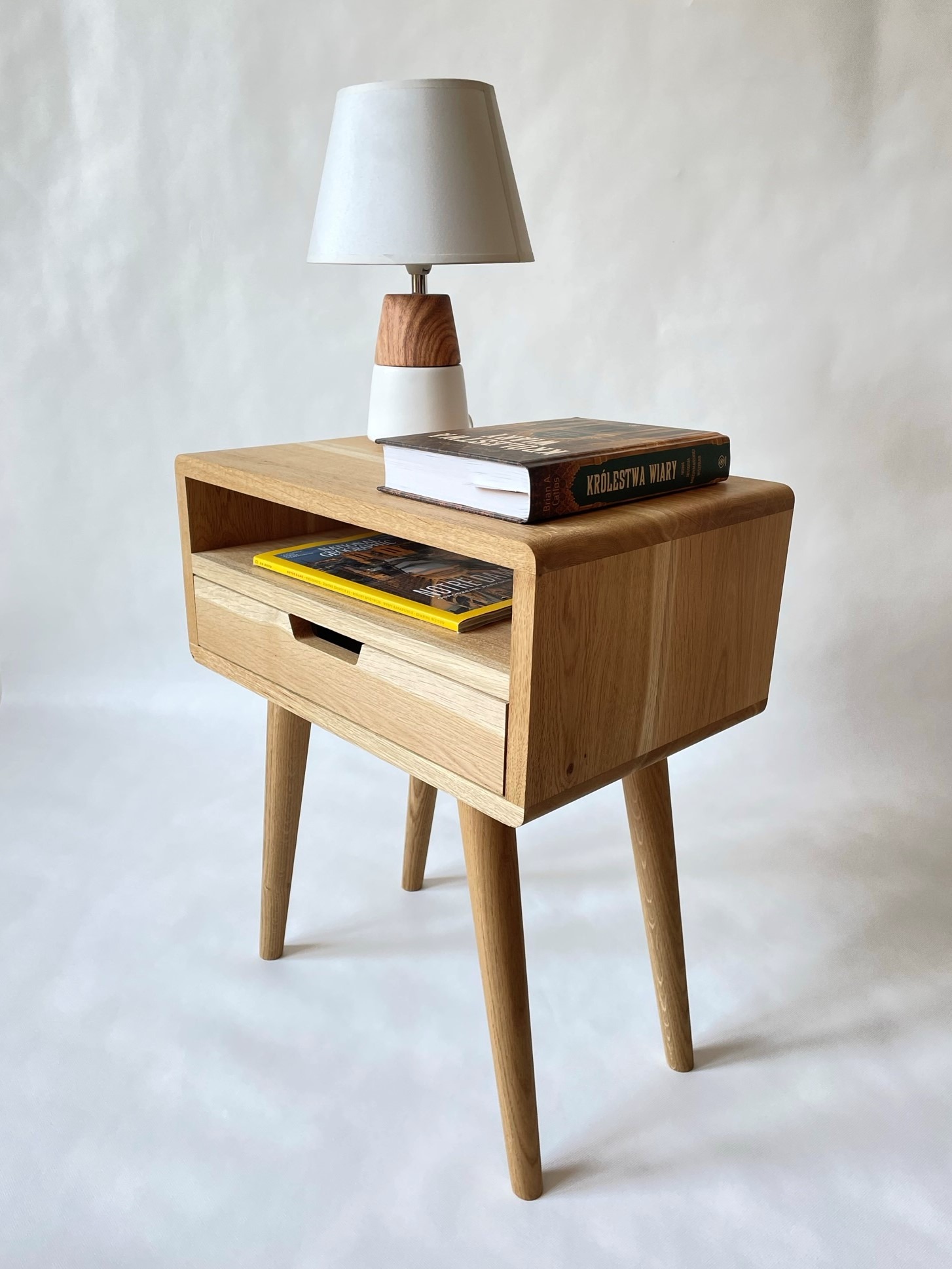 Drewniana szafka nocna w stylu retro z zaokrąglonymi krawędziami, na niej lampka nocna i książka 'Królestwa Wiary', a w otwartej półce kolorowy magazyn.