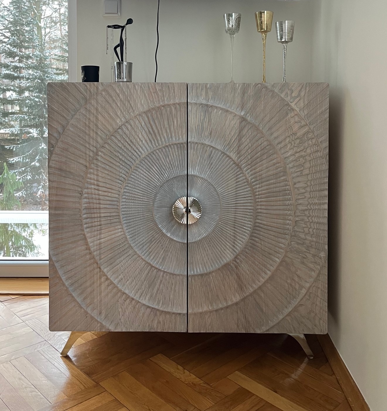 Dwudrzwiowa komoda z jasnego drewna z dekoracyjnym, okrągłym wzorem i srebrnymi uchwytami, stojąca na drewnianej podłodze w jodełkę, z widokiem na zaśnieżone drzewa za oknem.