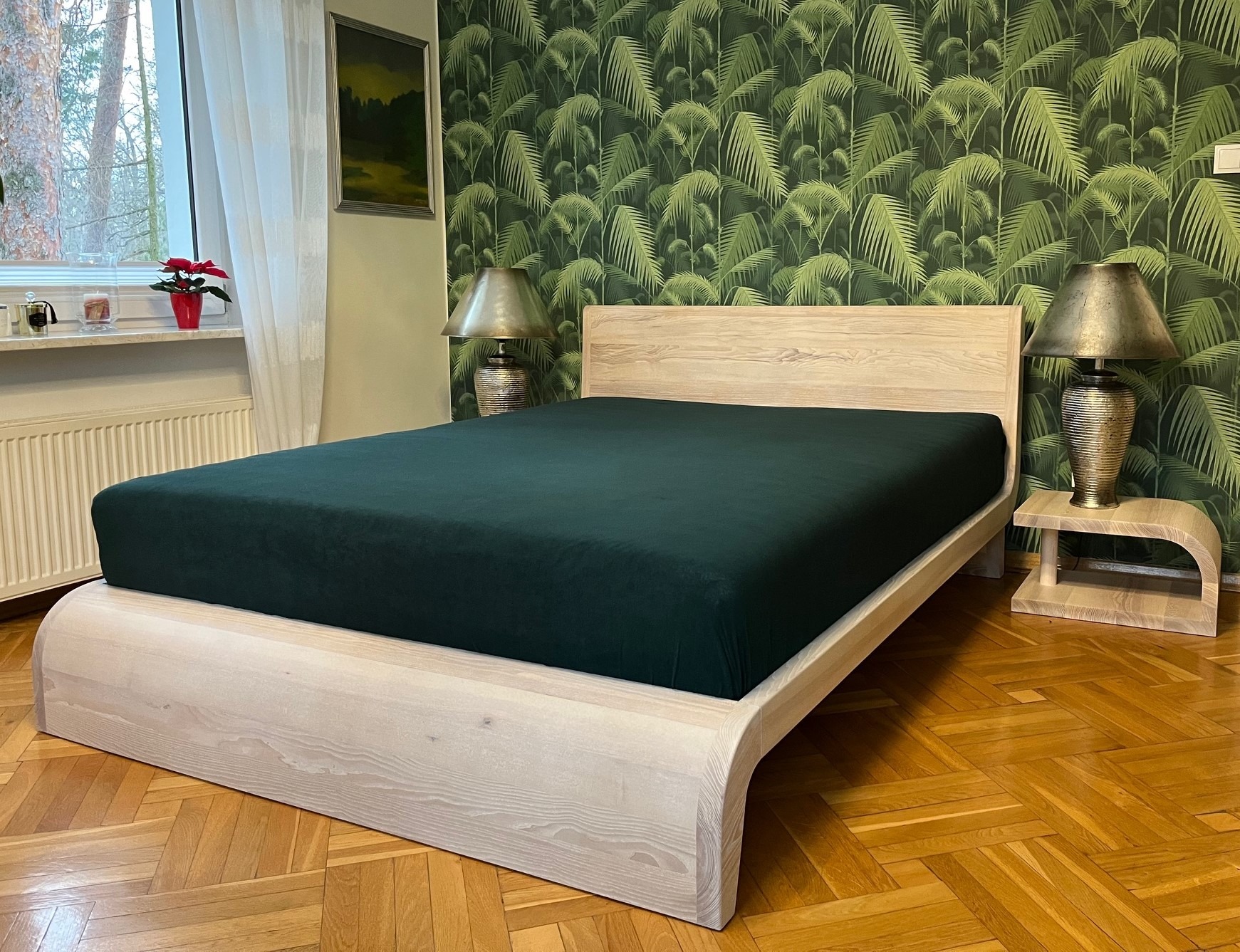 Drewniane łóżko z zaokrąglonymi krawędziami i zielonym prześcieradłem na tle ściany w tropikalne liście, z widocznymi lampkami nocnymi na stolikach.