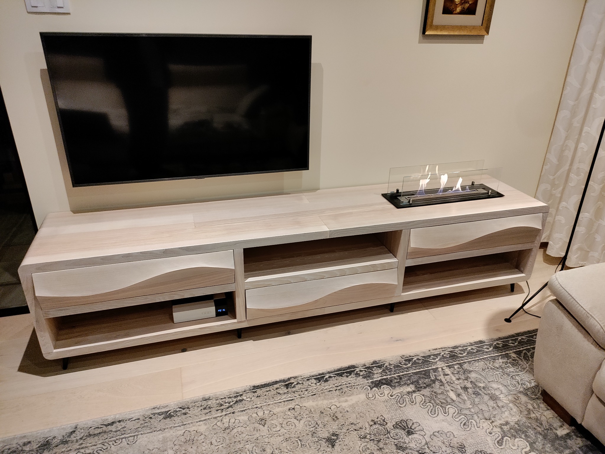 Jasna szafka RTV z drewnianym blatem i bio-kominkiem, z falistymi frontami szuflad, na tle ściany z telewizorem i dywanem.