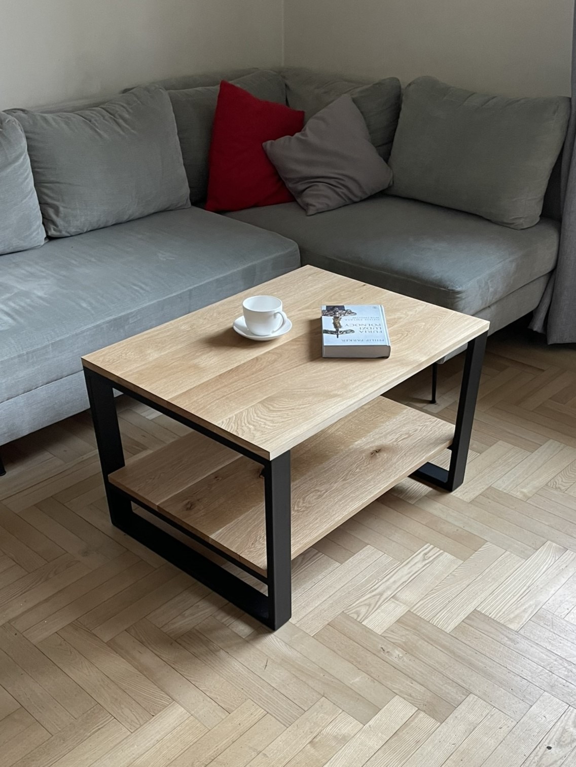 Stolik kawowy z jasnego drewna z czarnymi metalowymi nogami, z półką, na parkiecie ułożonym w jodełkę, obok szarej kanapy z poduszkami, na blacie biała filiżanka i książka.