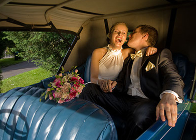 Uśmiechnięta panna młoda w białej sukni i pan młody w smokingu z muszką, siedzący w błękitnym, zabytkowym samochodzie z bukietem ślubnym na siedzeniu.