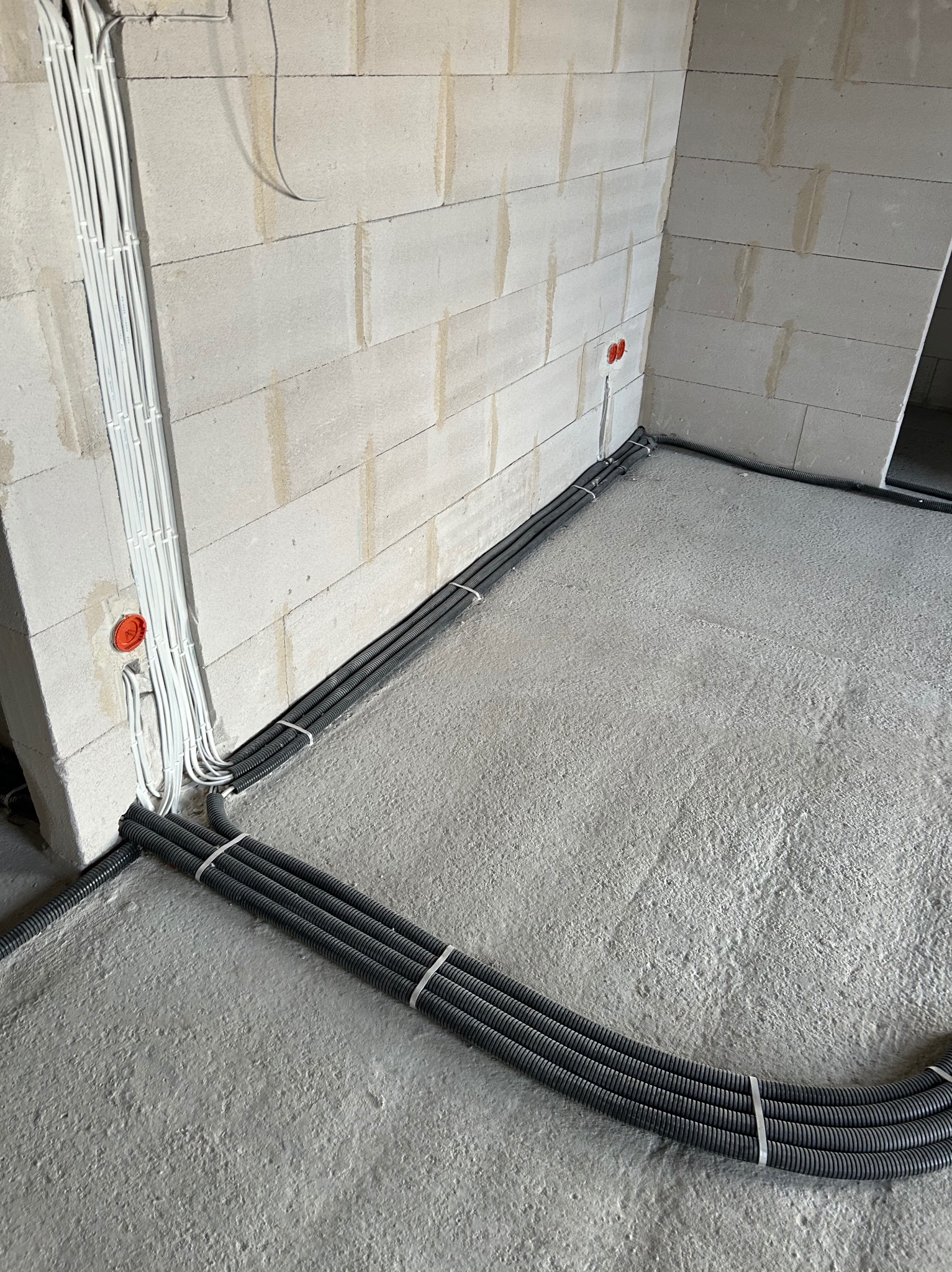 Szare rury instalacji elektrycznej ułożone na szarej podłodze, przymocowane do ściany z białych pustaków. Widoczne pomarańczowe puszki elektryczne.