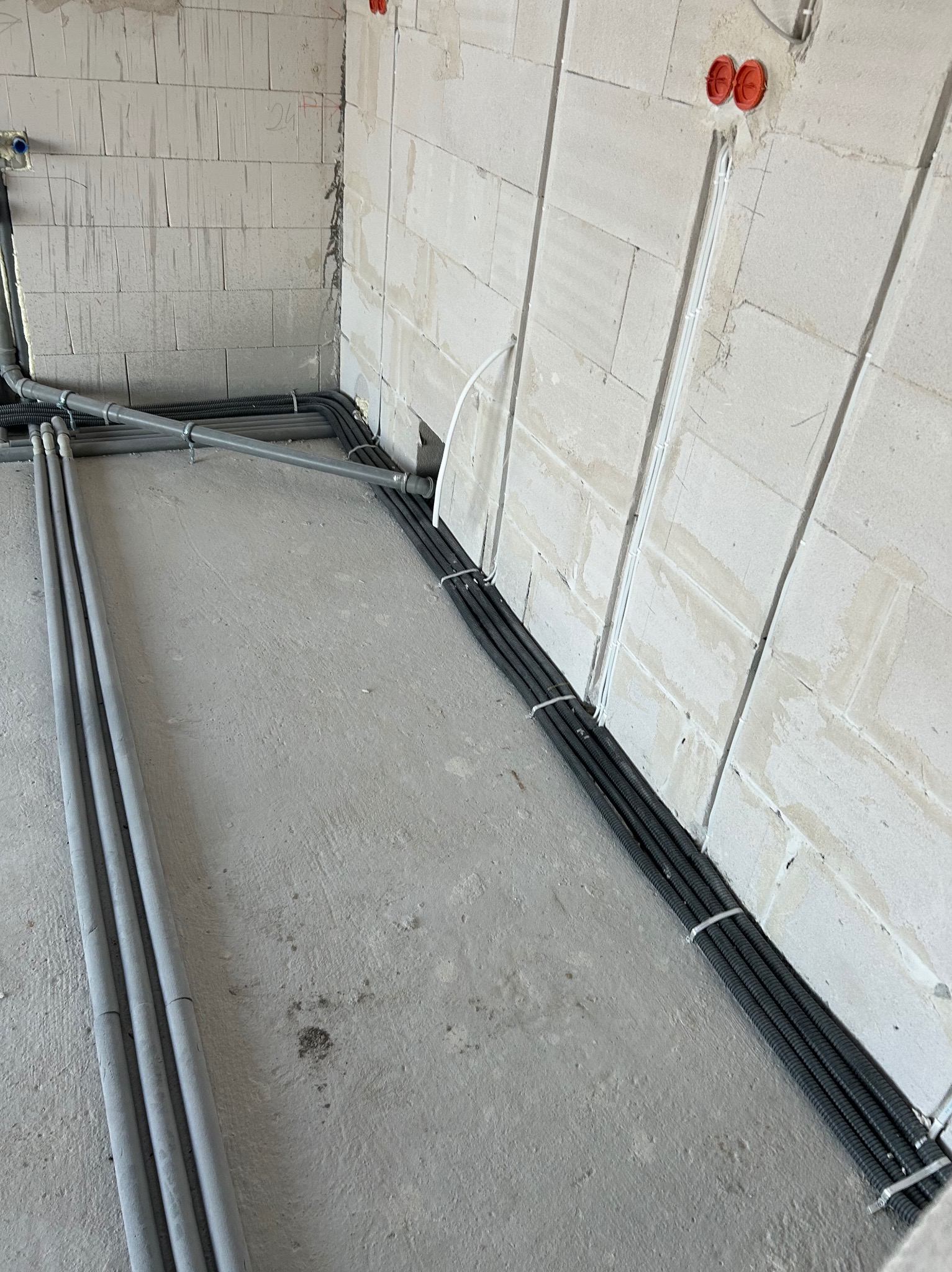 Szare i czarne rury prowadzone po podłodze i ścianie z betonu komórkowego. Widoczne puszki elektryczne na ścianie.