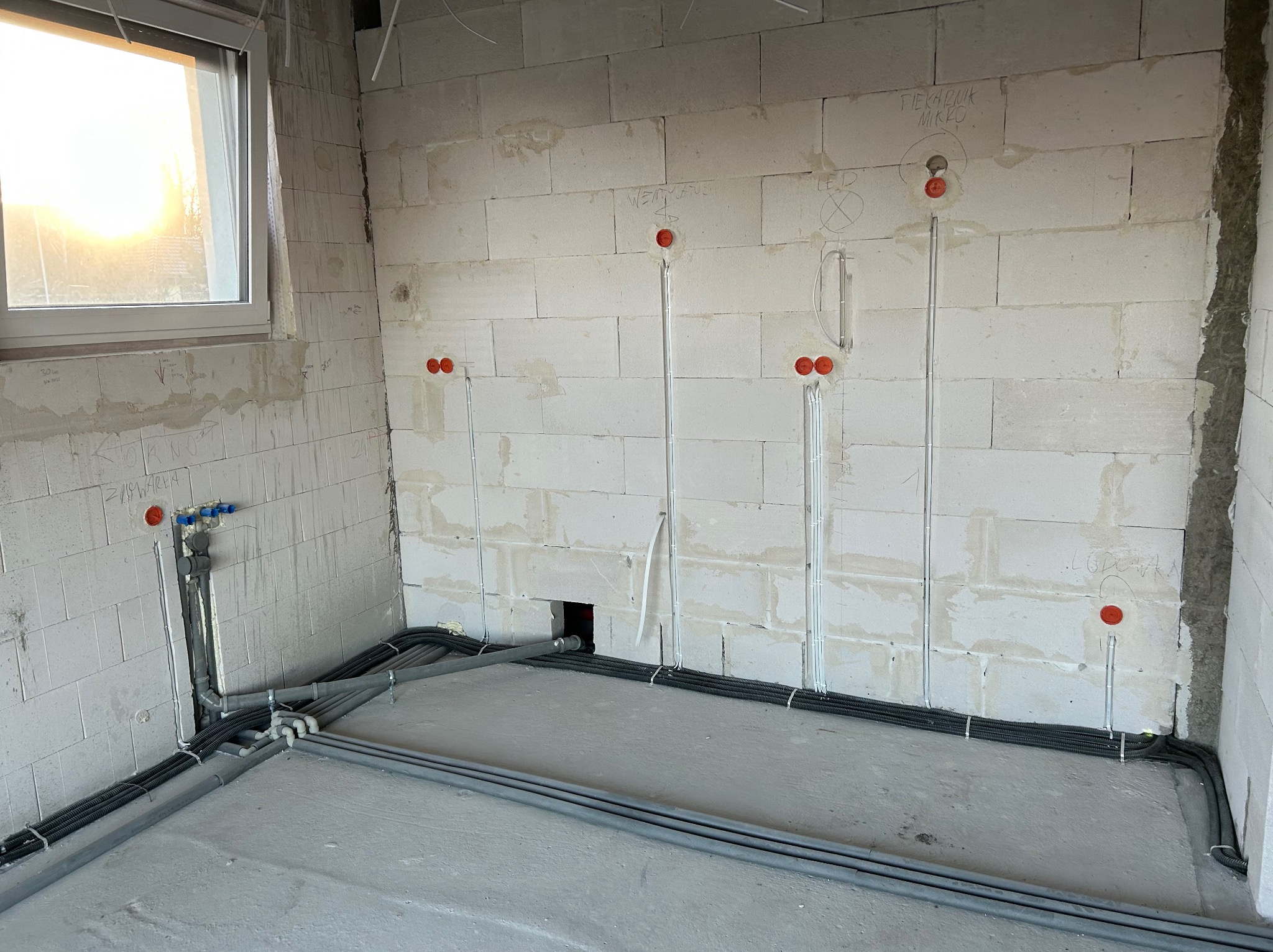 Instalacja elektryczna w surowym wnętrzu: widoczne kable w peszlach ułożone wzdłuż ścian, puszki instalacyjne z pomarańczowymi dekielkami, okno z widokiem na zachód słońca.