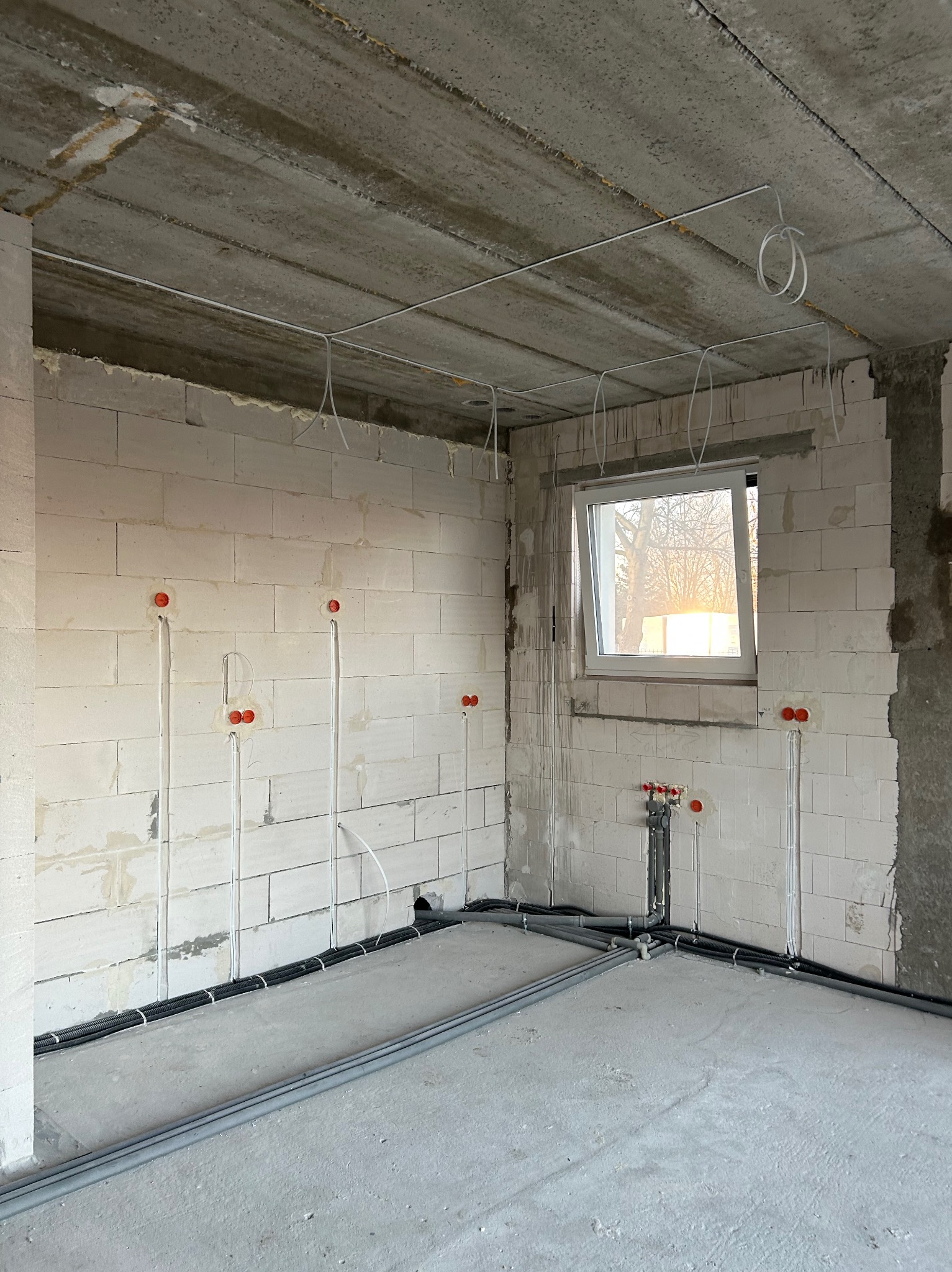 Surowy stan: ściany z betonu komórkowego z puszkami i przewodami elektrycznymi, rury na podłodze, okno z widokiem na drzewa w tle. Wnętrze w trakcie prac instalacyjnych.