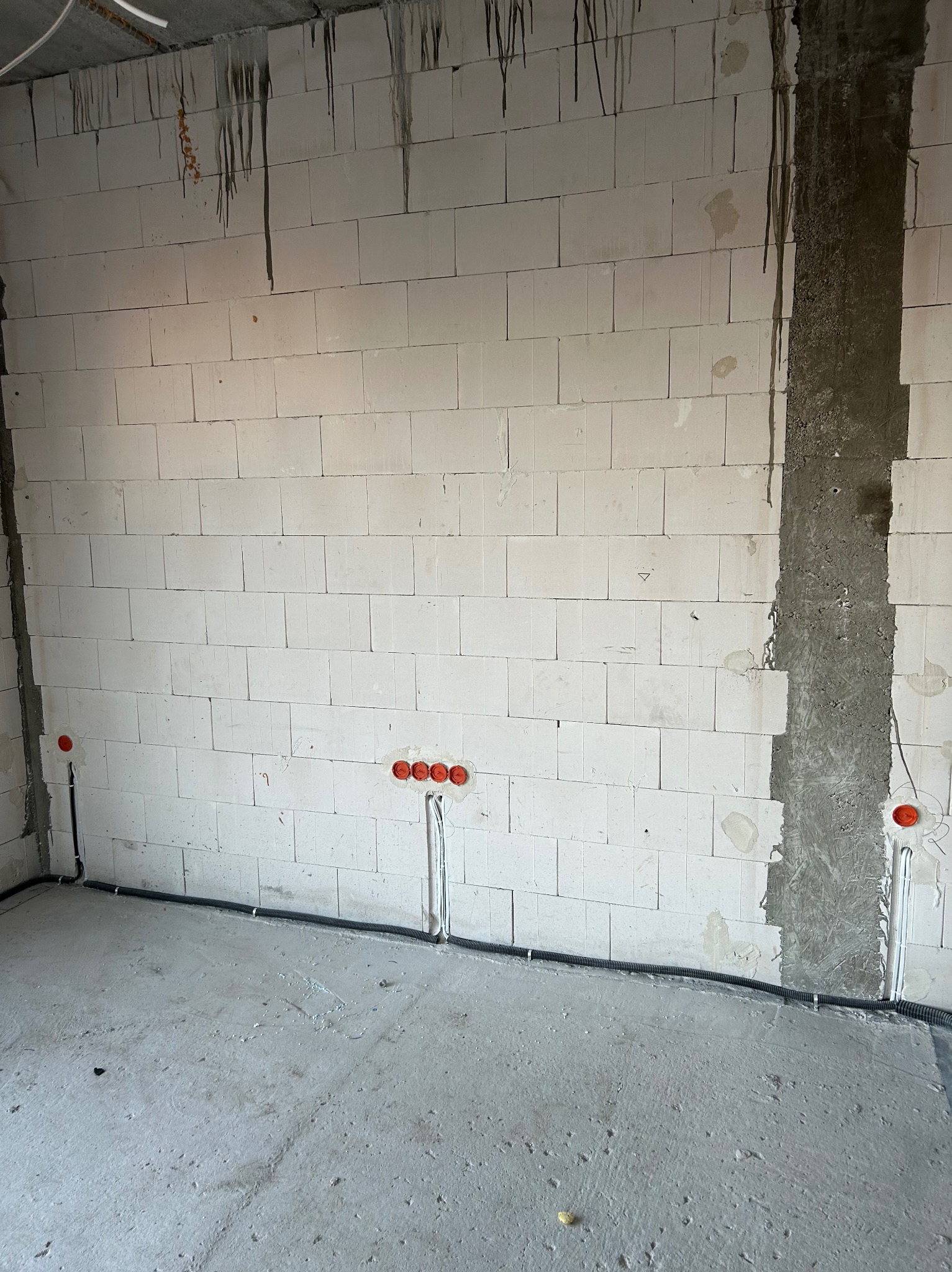 Wnętrze w trakcie prac: ściana z białego bloczka z widocznymi puszkami i przewodami elektrycznymi, czarna rura karbowana wzdłuż podłogi. Surowy stan wykończenia.