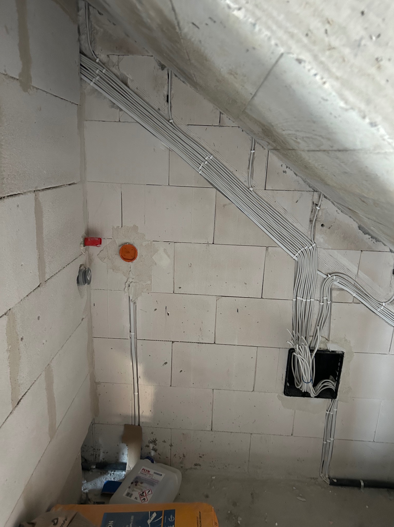 Szereg białych kabli elektrycznych w peszlach, przymocowanych do białej ściany z pustaków. Widoczne puszki instalacyjne i fragment poddasza.