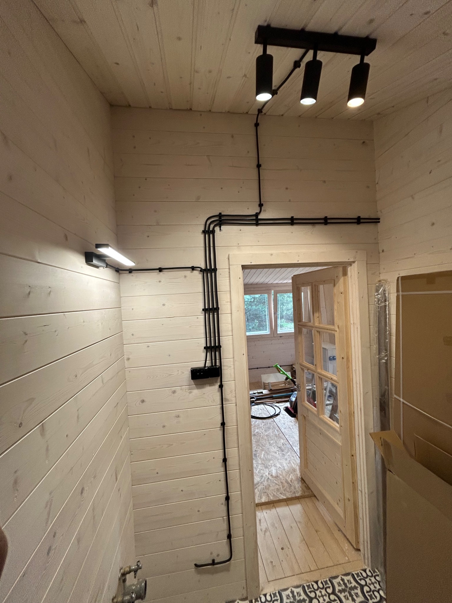 Korytarz z drewnianą boazerią i nową instalacją elektryczną natynkową w czarnych rurkach. Widoczne lampy sufitowe, kinkiet i gniazdka. Otwarta drewniana rama drzwiowa.