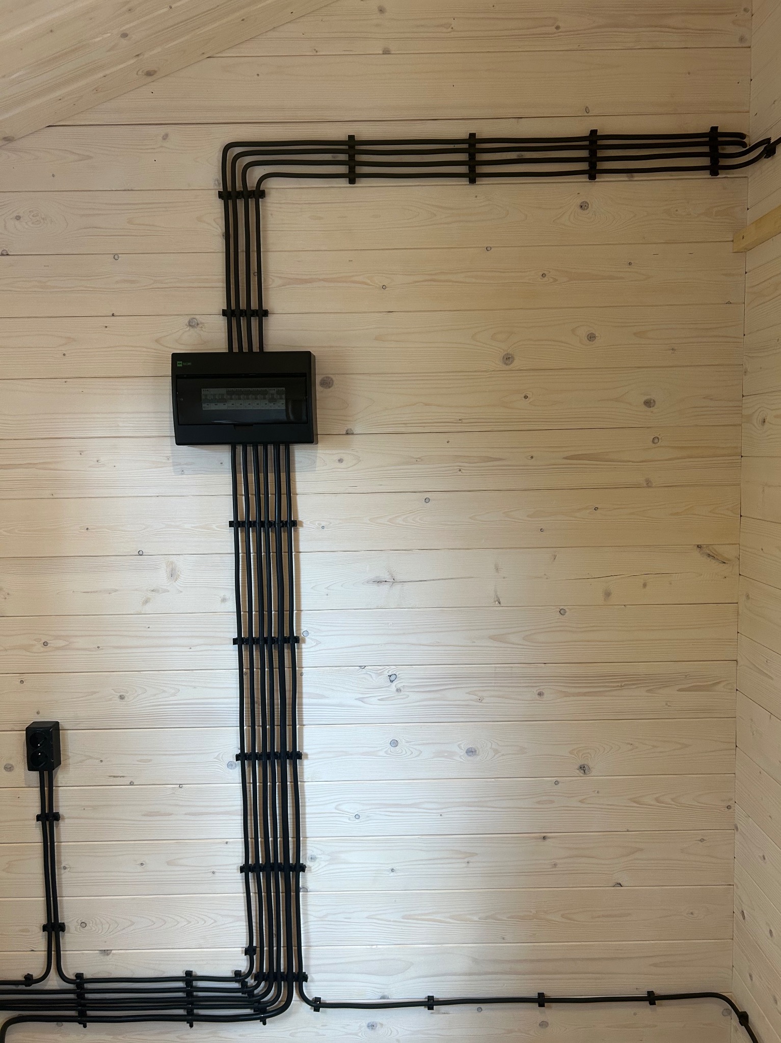 Czarna instalacja elektryczna natynkowa, z puszką i gniazdkiem, zamocowana na jasnej, drewnianej ścianie wewnątrz budynku. Estetyczne ułożenie przewodów.