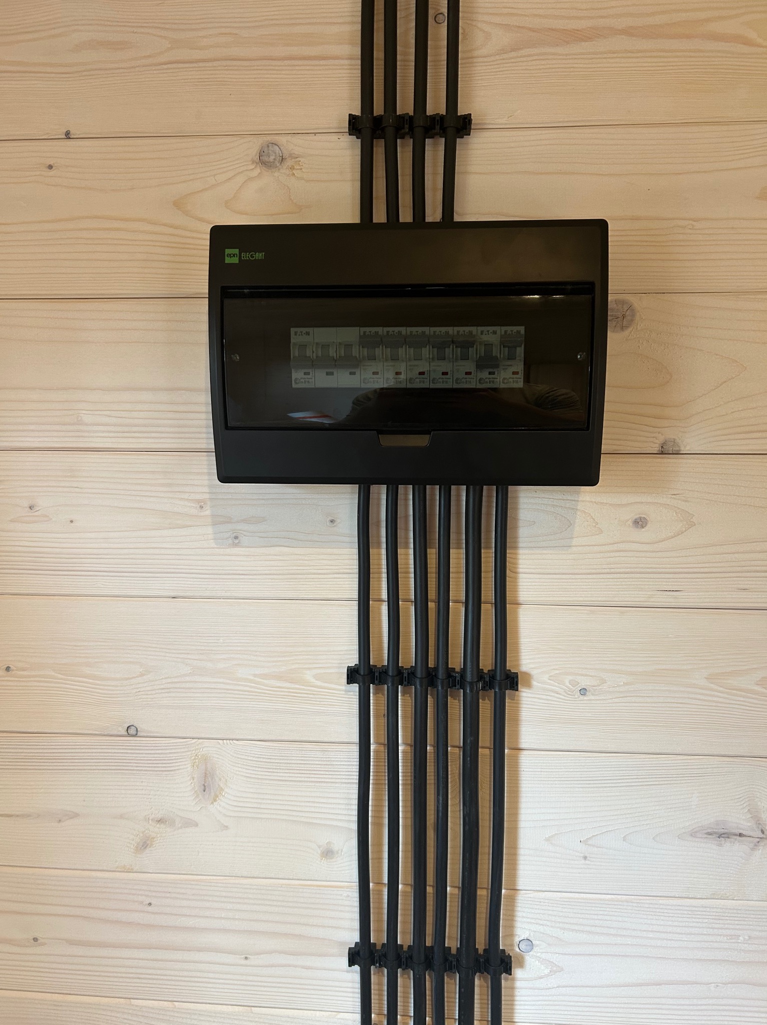 Czarna skrzynka rozdzielcza Eaton Elegant na jasnej, drewnianej ścianie. Kable prowadzone pionowo w dół, przymocowane uchwytami. Widoczne bezpieczniki wewnątrz.