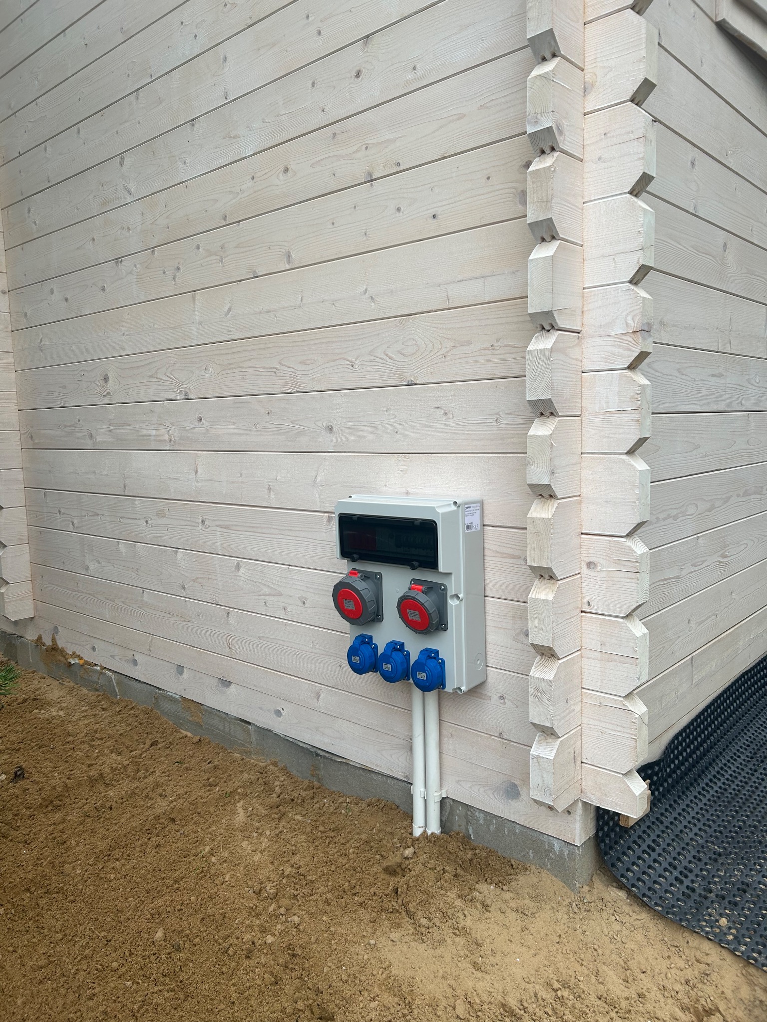 Szara skrzynka elektryczna z czerwonymi i niebieskimi gniazdami, zamontowana na ścianie z jasnego drewna. Widoczne rury doprowadzające kable.