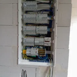 Rozdzielnica elektryczna z zainstalowanymi bezpiecznikami i przewodami, zamontowana w ścianie z betonu komórkowego, widoczne elementy instalacji elektrycznej oraz licznik energii.