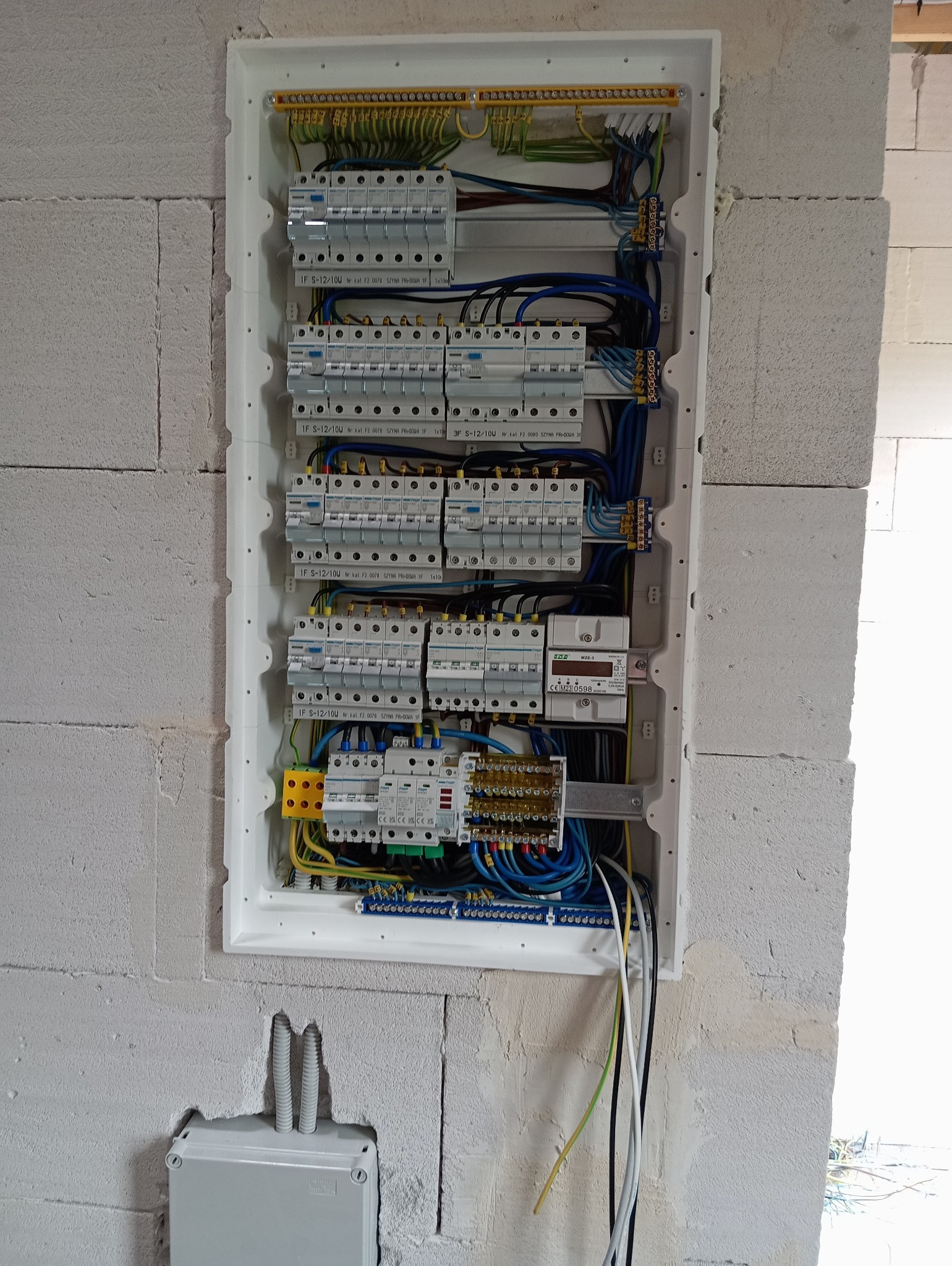 Rozdzielnica elektryczna z zainstalowanymi bezpiecznikami i przewodami, zamontowana w ścianie z betonu komórkowego, widoczne elementy instalacji elektrycznej oraz licznik energii.