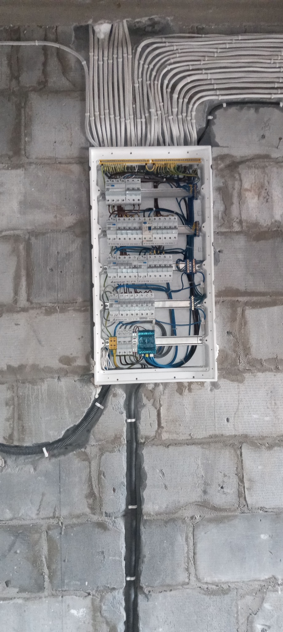 Otwarta skrzynka elektryczna z zamontowanymi bezpiecznikami i okablowaniem na surowej, betonowej ścianie z widocznymi przewodami w peszlach.
