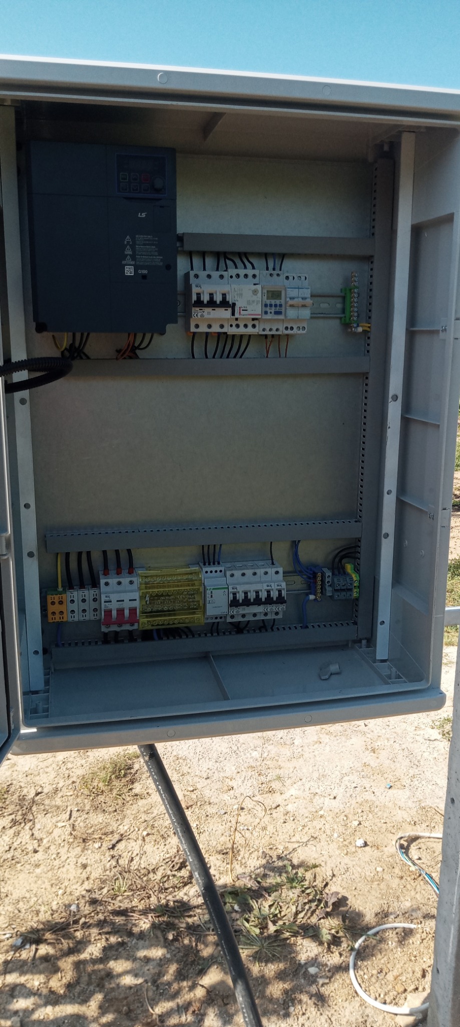 Otwarta szafa elektryczna na zewnątrz z widocznymi bezpiecznikami, przewodami i sterownikiem LS, umieszczona na tle ziemi i nieba.