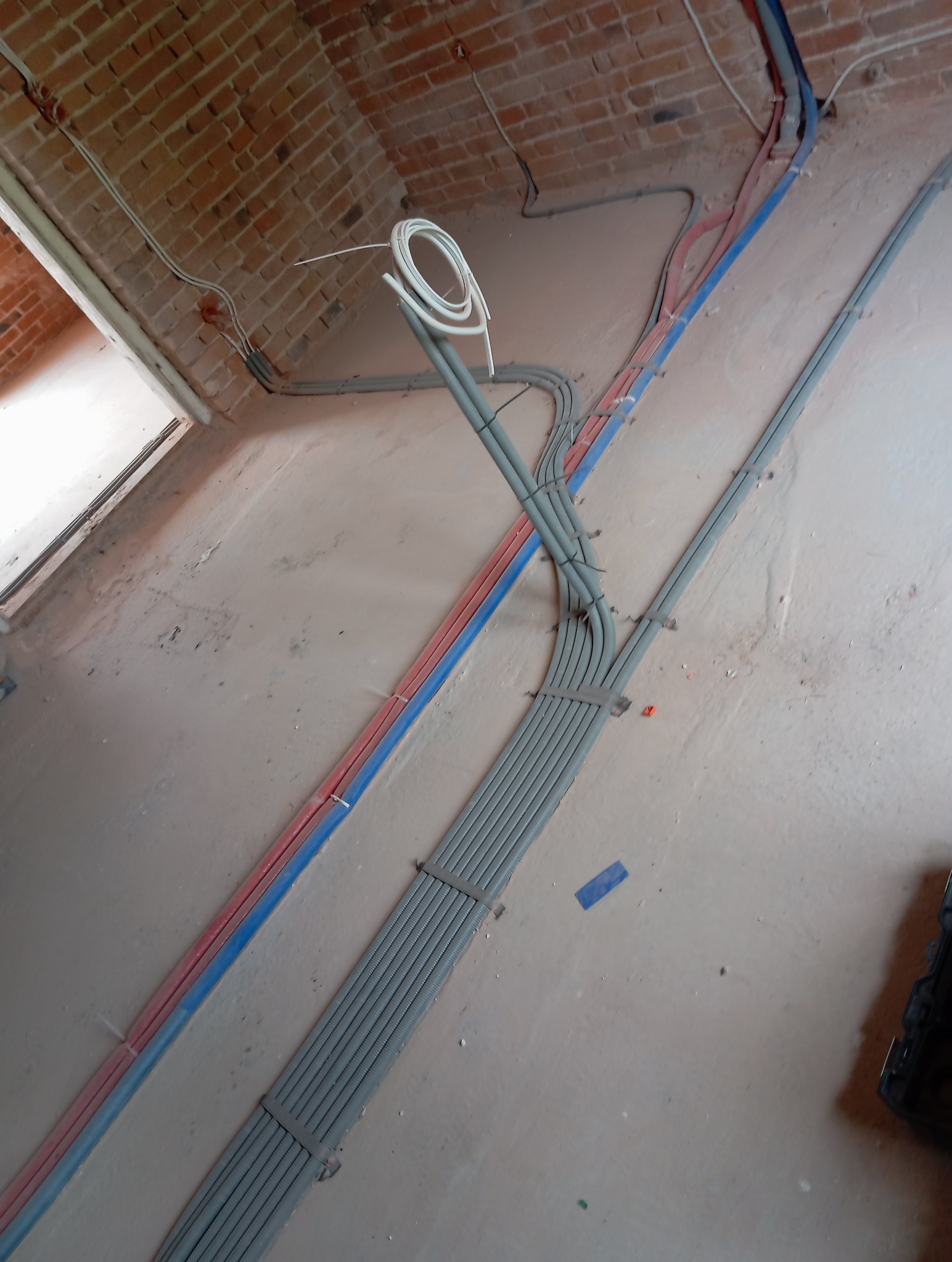 Szare rury instalacji w surowym wnętrzu, widok z góry. Instalacje prowadzone w Jastrzębskiej Woli przed wylaniem posadzki. Czerwone i niebieskie rury w tle.