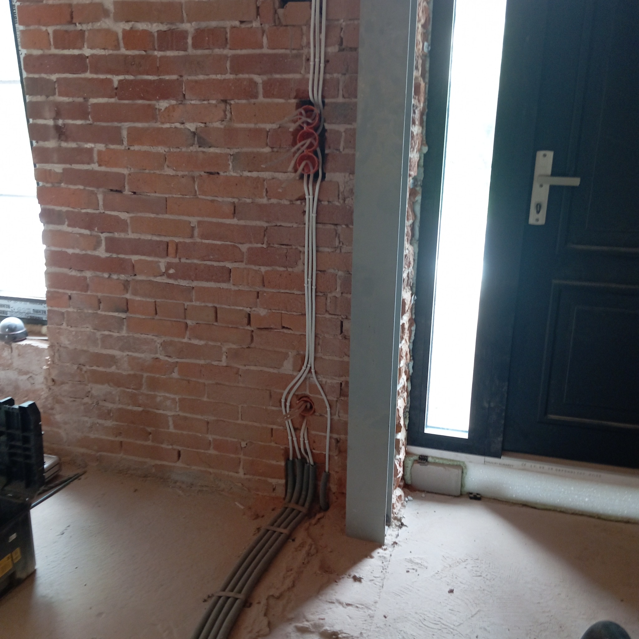 Instalacja elektryczna w trakcie montażu na ceglanej ścianie, widoczne kable w peszlach prowadzone do otworów w ścianie. Obok framuga drzwi.