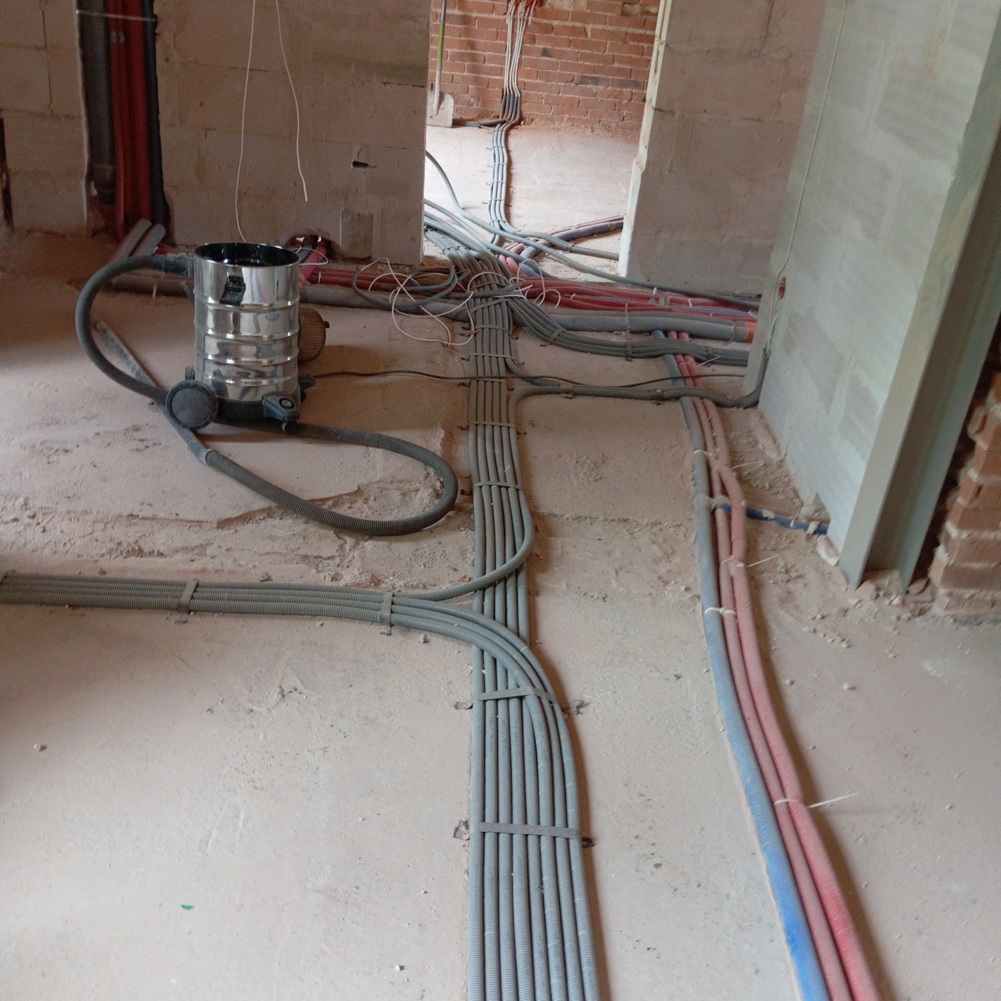 Szare i kolorowe rury instalacyjne ułożone na podłodze w surowym stanie budynku. Widoczny odkurzacz przemysłowy i fragmenty ścian z cegły.