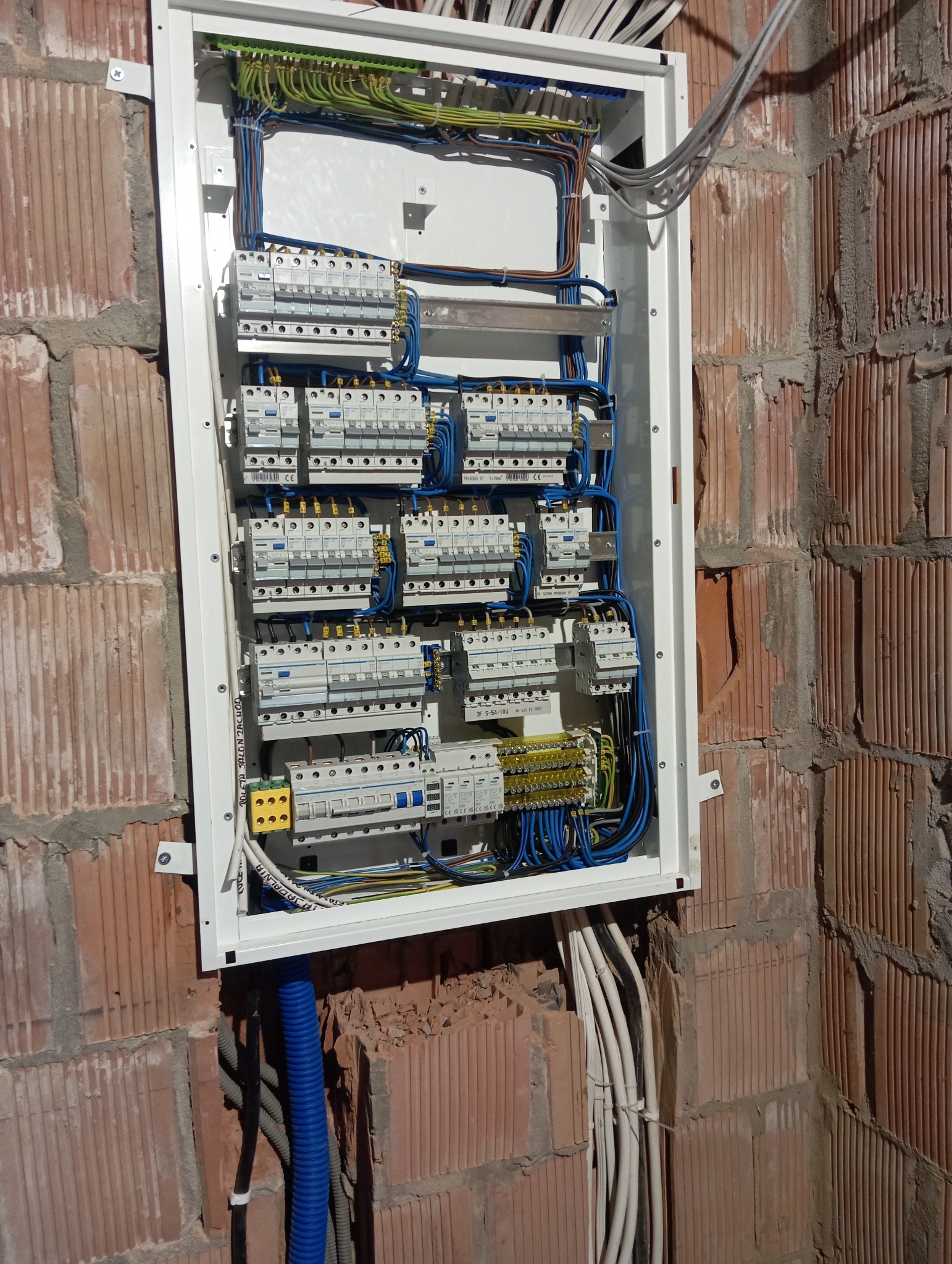 Rozdzielnica elektryczna z bezpiecznikami i okablowaniem, zamontowana na surowej ścianie z cegły. Widoczne kable w peszlach.