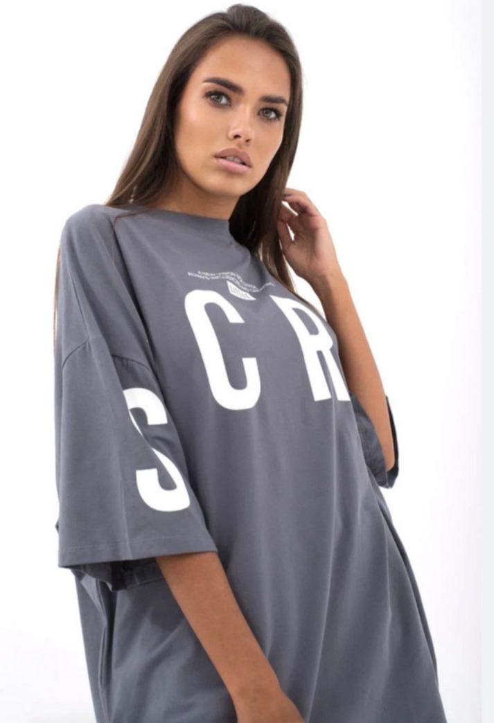 Kobieta pozuje w szarej, oversize'owej koszulce z białym napisem 'SCR' i drobnym tekstem powyżej, z ręką uniesioną do włosów.