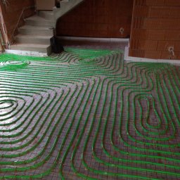 Szymon Michaluk - Zielone rury ogrzewania podłogowego ułożone spiralnie na szarej macie z czerwoną siatką w surowym wnętrzu, widoczne schody i fragment ściany z pustaków.