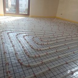 Szymon Michaluk - Białe rury ogrzewania podłogowego z czerwonymi uchwytami mocowane na folii z nadrukiem kratki, widok z góry, w tle okno i fragment ściany.