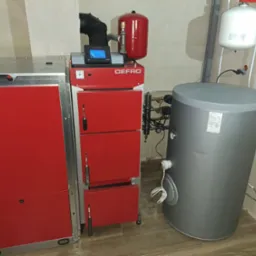 Czerwony piec CO marki Defro z zasobnikiem i zbiornikiem wyrównawczym w kotłowni, widoczne elementy instalacji.