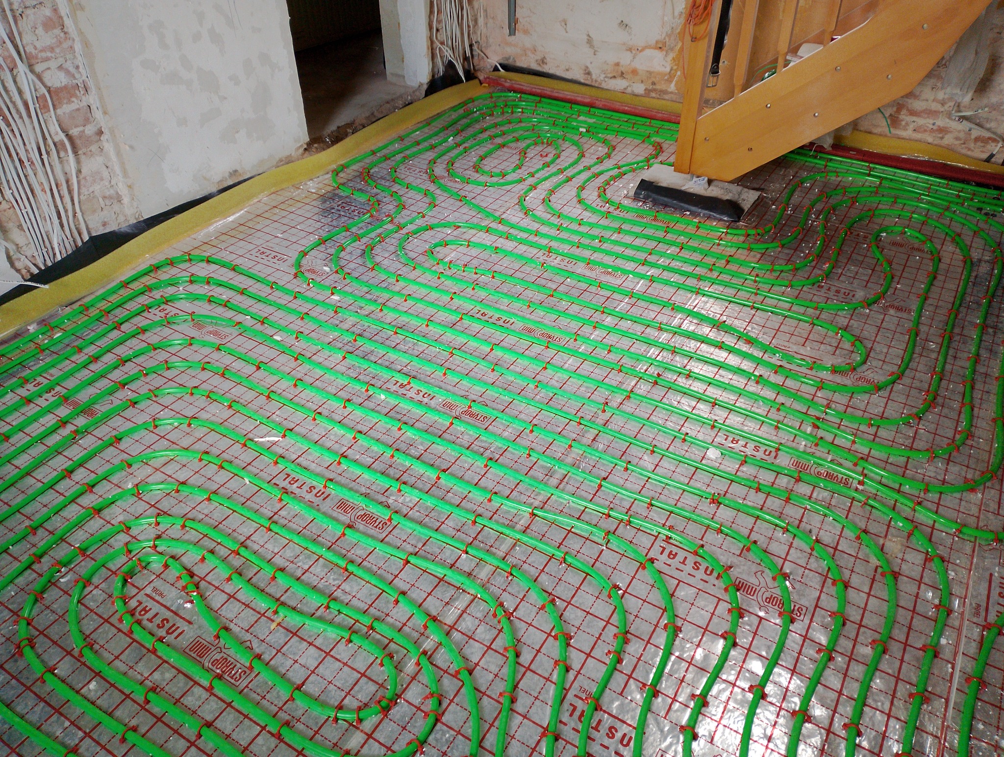 Zielone rurki ogrzewania podłogowego ułożone na folii z siatką, widoczne fragmenty ścian i schodów w surowym stanie.