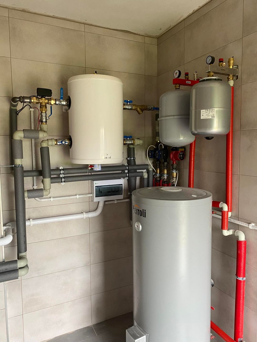 Zestaw urządzeń grzewczych: dwa zbiorniki buforowe (biały i szary Ferroli), dwa naczynia przeponowe, rozdzielacz z manometrami, orurowanie w izolacji, zamontowane na ścianie z płytek ceramicznych.