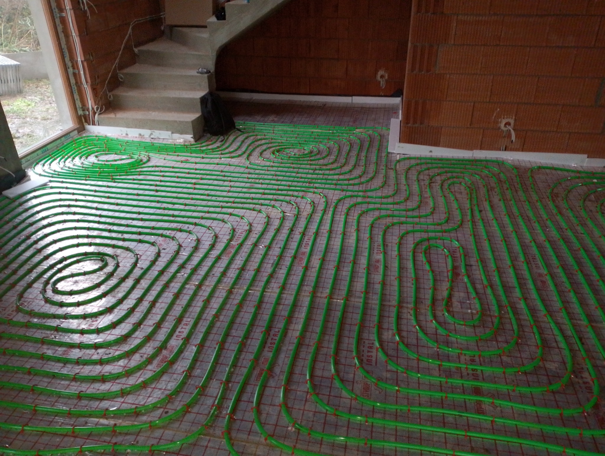 Zielone rury ogrzewania podłogowego ułożone spiralnie na szarej macie z czerwoną siatką w surowym wnętrzu, widoczne schody i fragment ściany z pustaków.