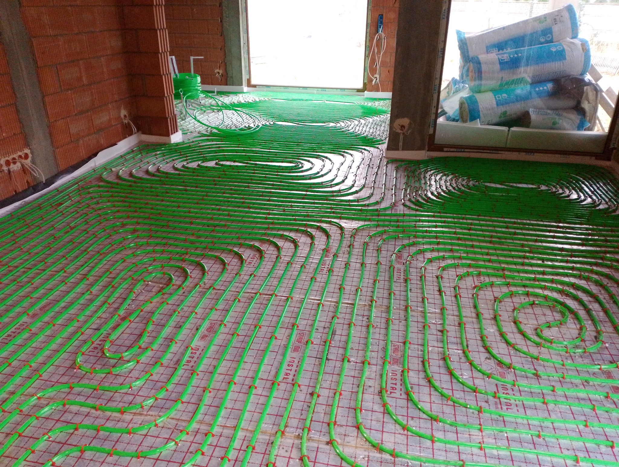 Układanie zielonych rurek ogrzewania podłogowego na siatce montażowej w surowym stanie budynku, widoczne pakiety izolacji termicznej przy oknie.