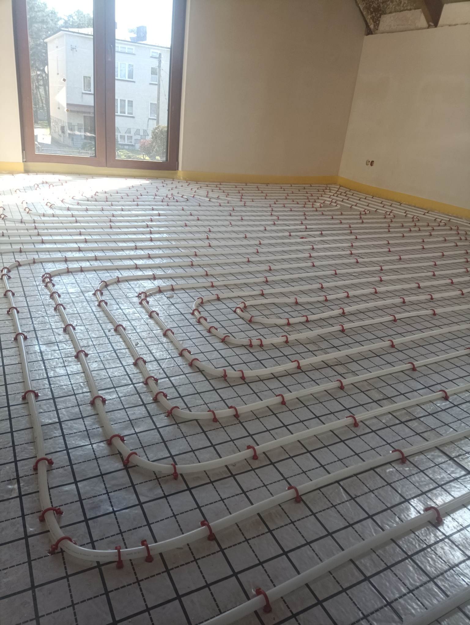 Białe rury ogrzewania podłogowego z czerwonymi uchwytami mocowane na folii z nadrukiem kratki, widok z góry, w tle okno i fragment ściany.