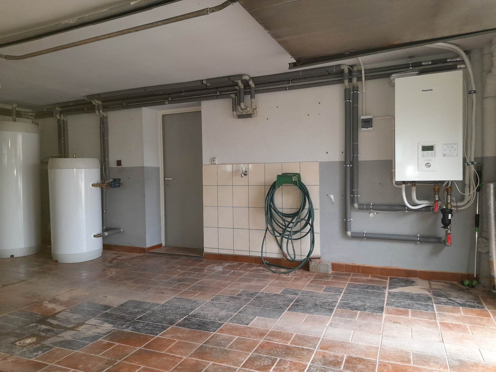 Piwnica z dwoma białymi zbiornikami, piecem gazowym Panasonic i szarymi rurami grzewczymi na ścianach, zielony wąż ogrodowy zwinięty na wieszaku, podłoga wyłożona płytkami w odcieniach brązu...