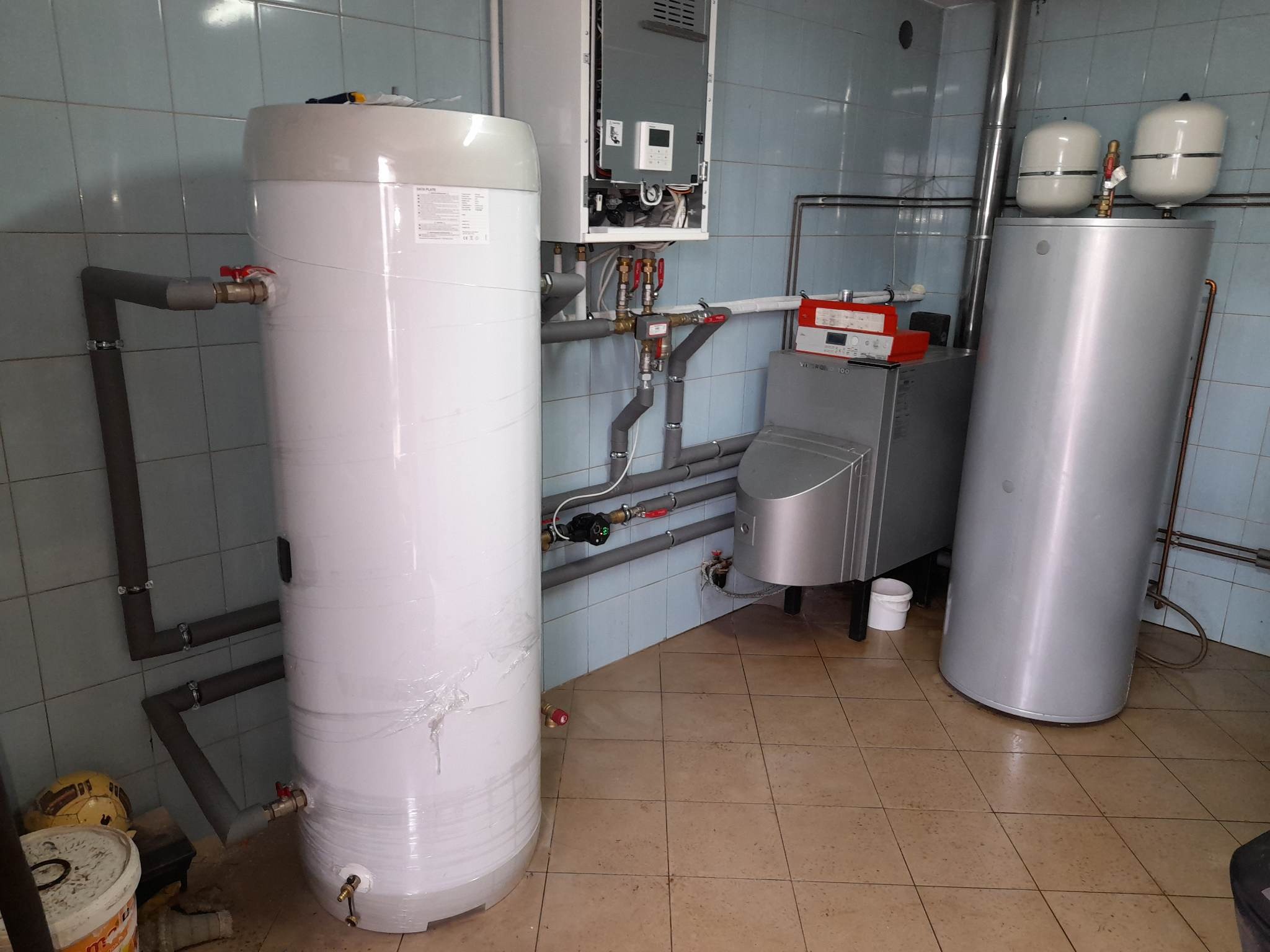Kotłownia z białym zasobnikiem ciepłej wody owiniętym folią, piecem gazowym i dwoma zbiornikami wyrównawczymi na tle niebieskich płytek ceramicznych.