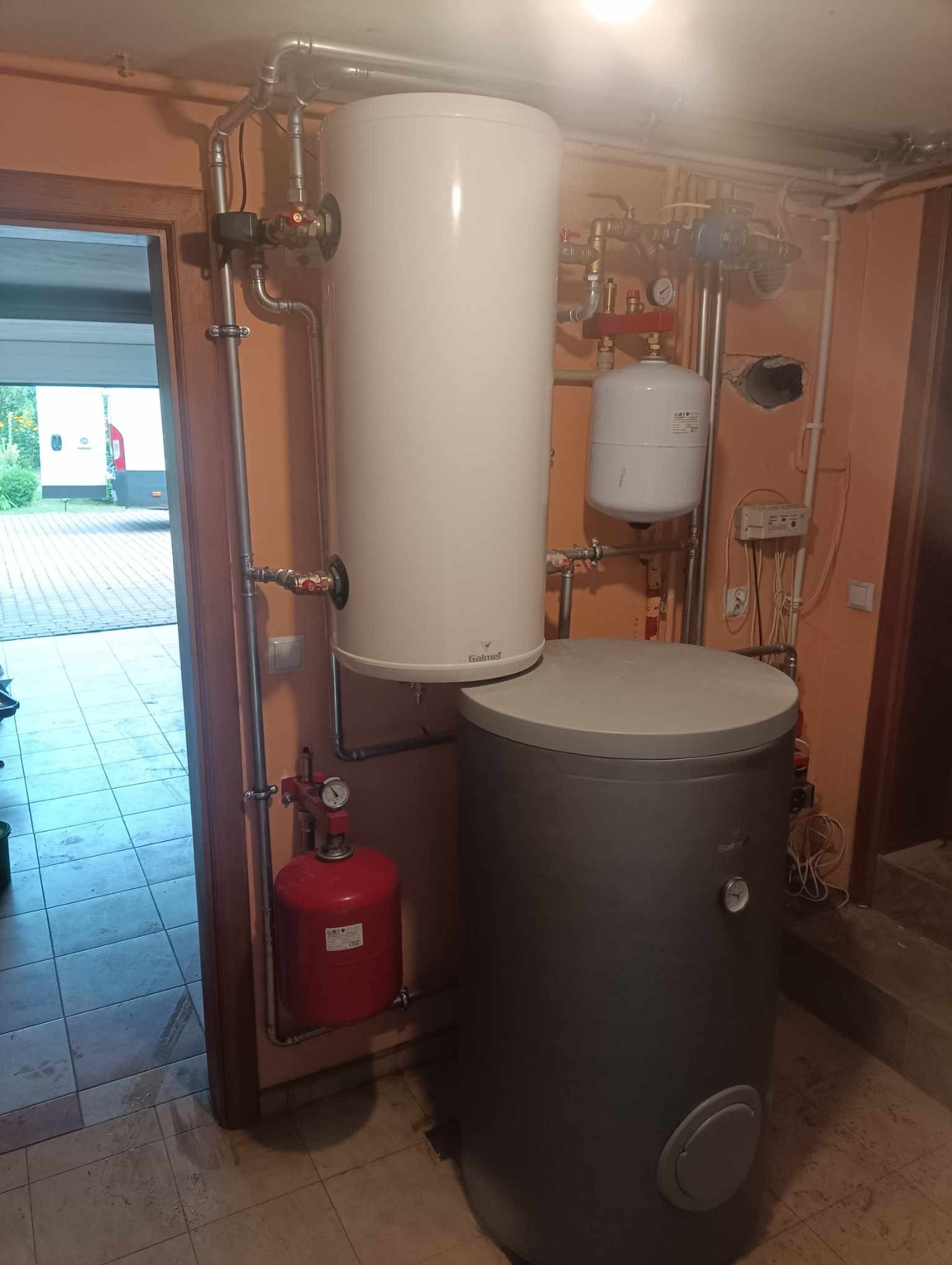 Biały zasobnik ciepłej wody użytkowej Galmet i szary bufor ciepła z podłączeniami hydraulicznymi i elektrycznymi, widoczne w piwnicy domu.